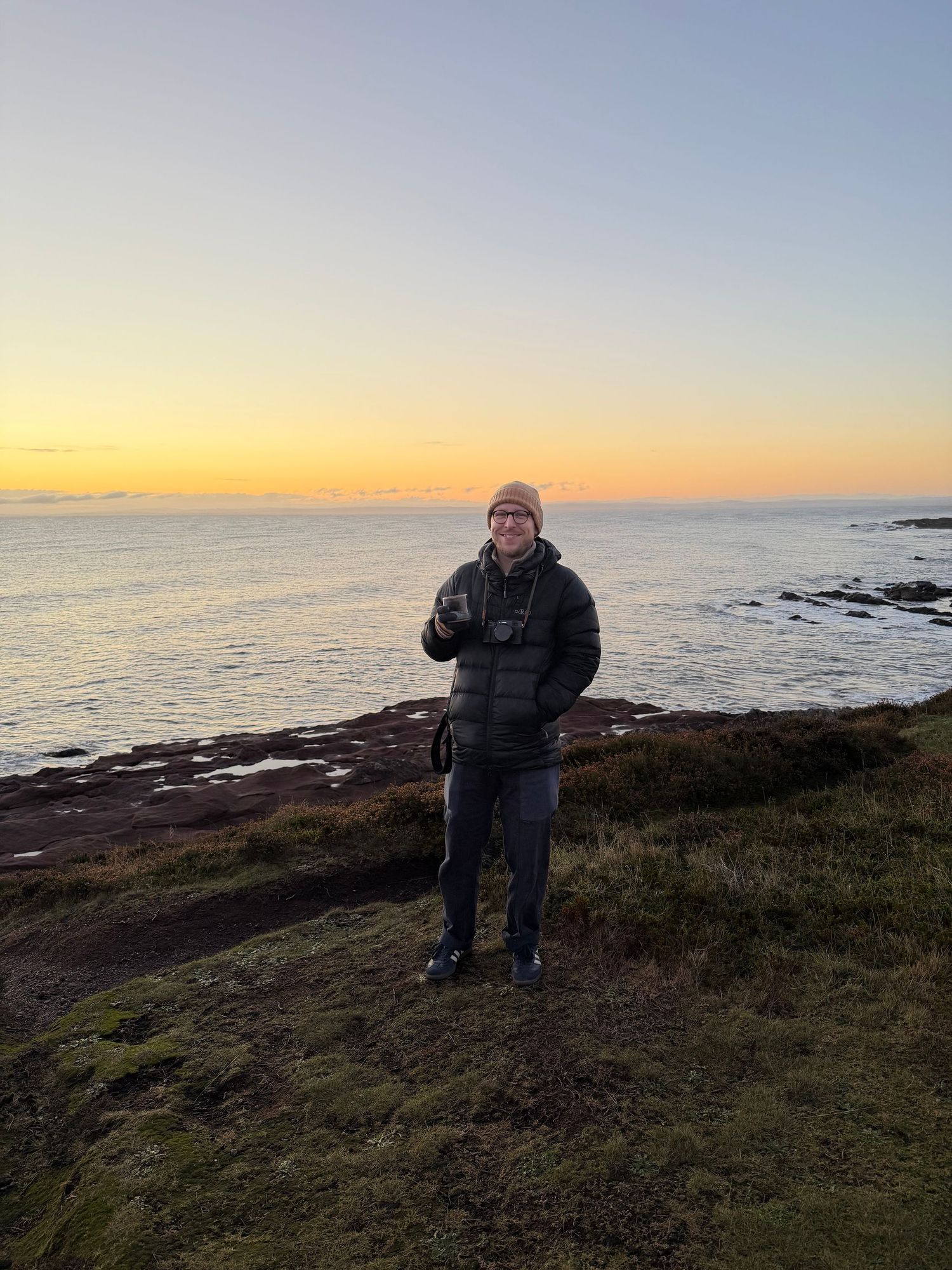 Person standing on rocky coastal cliffs at golden hour sunset, wearing puffer jacket and beanie, holding a drink.
