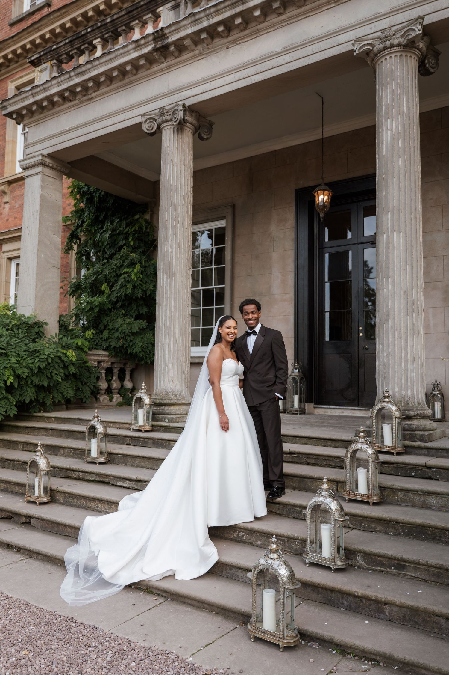 A couple in wedding attire poses on the grand steps of a historic building with classic columns and lanterns.