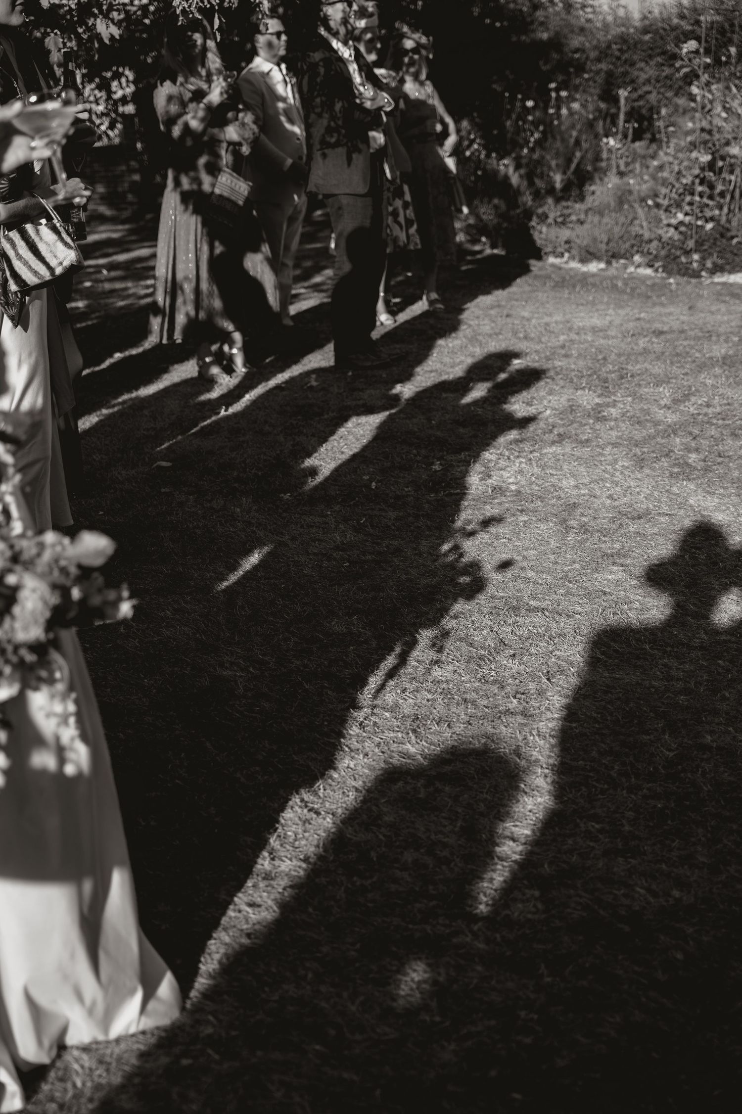 A black and white photograph shows long shadows cast by a crowd of people standing on grass in outdoor lighting.