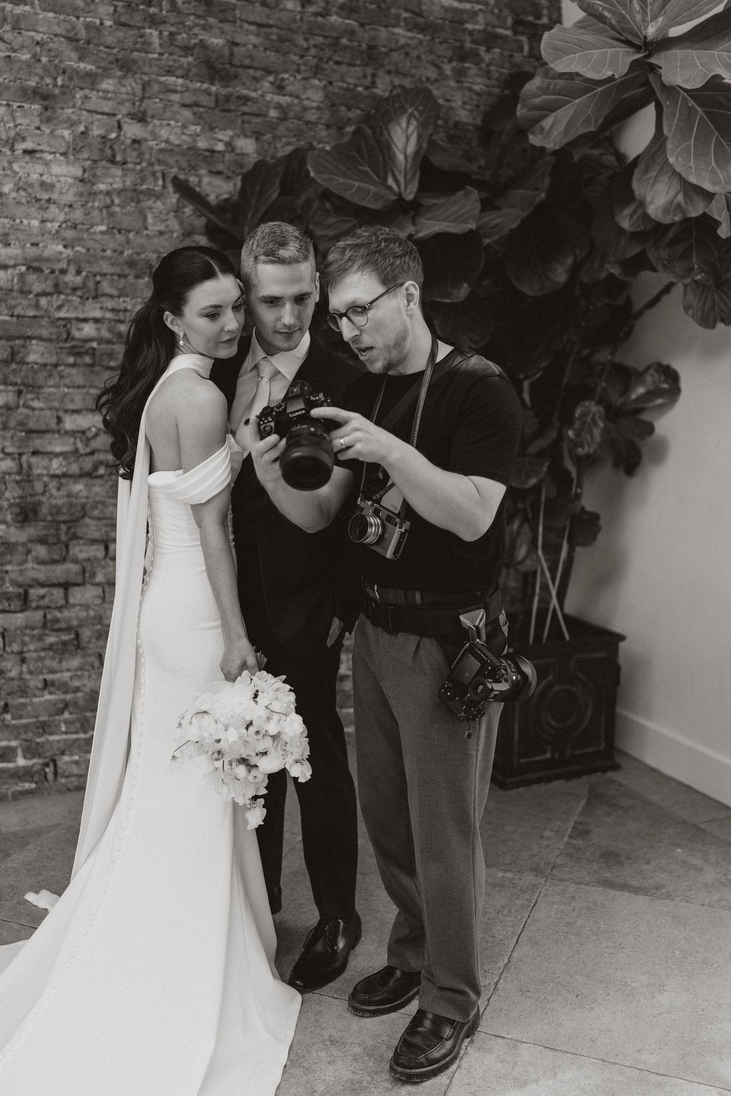 Black and white photo of a wedding photographer showing camera preview to bride and groom near brick wall.