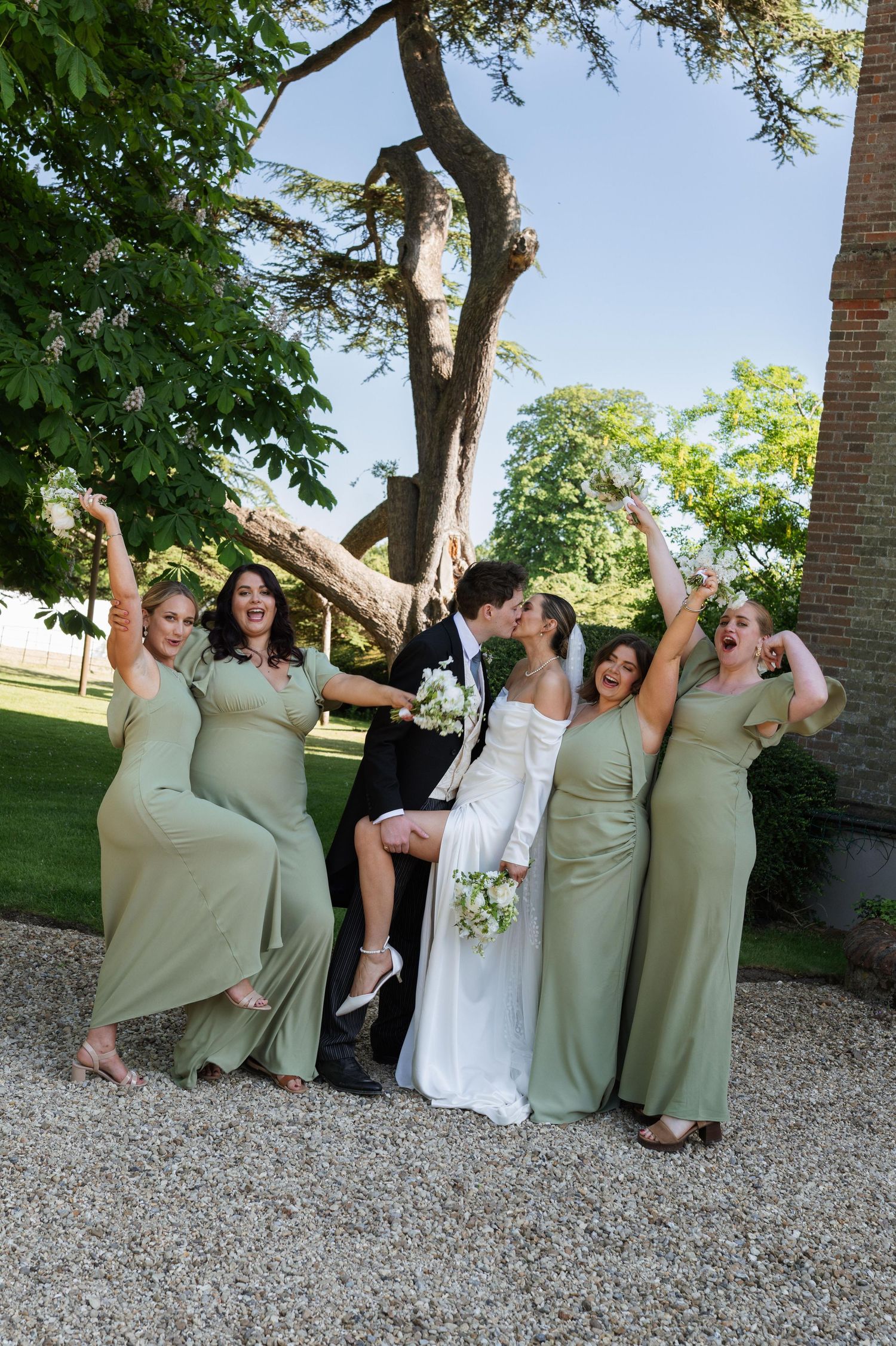 Wedding party in sage green dresses poses playfully under a tree on a gravel path.