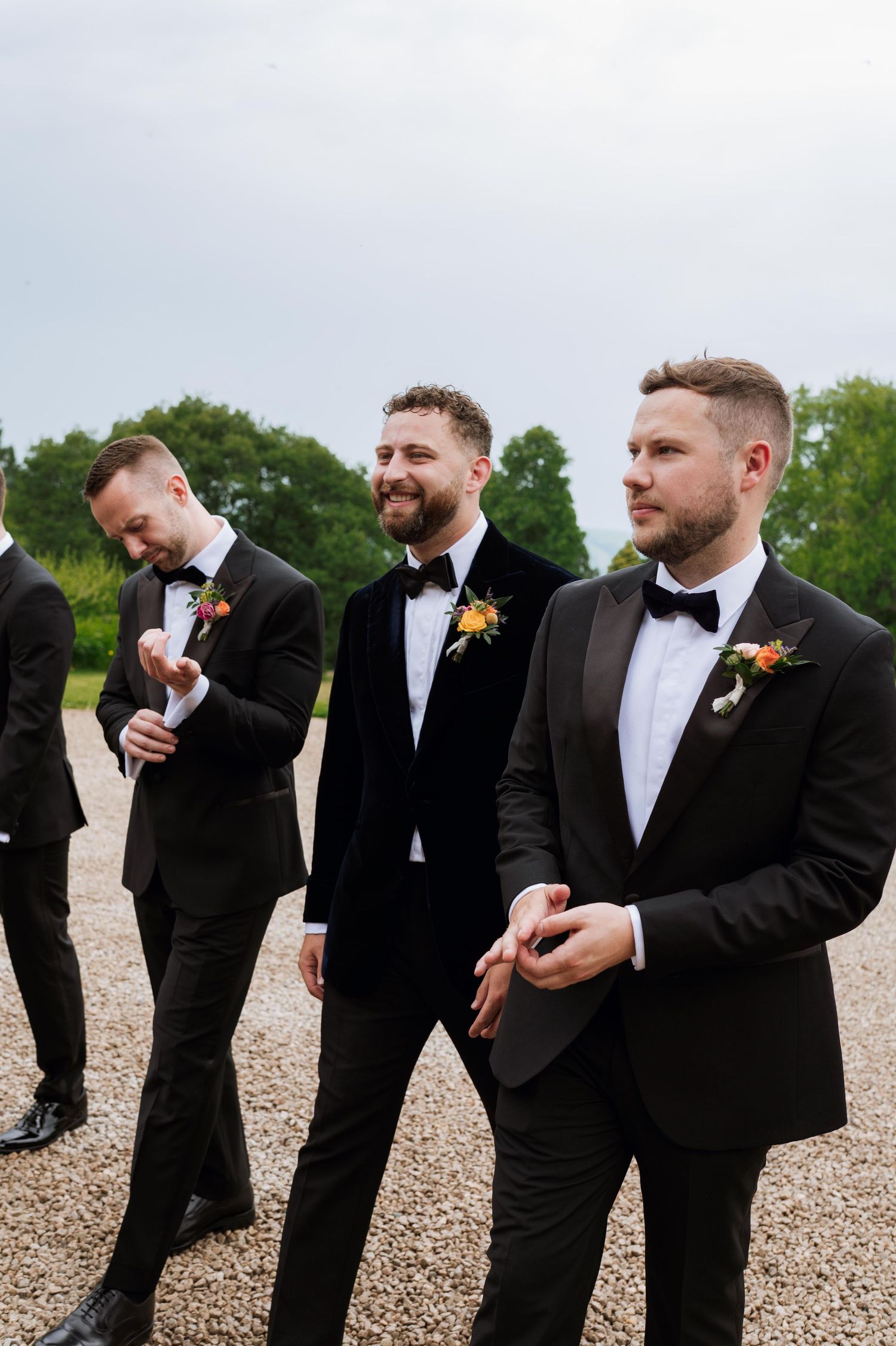 Groomsmen in black tuxedos and bow ties stand together on a gravel path while sharing a candid moment at a wedding.