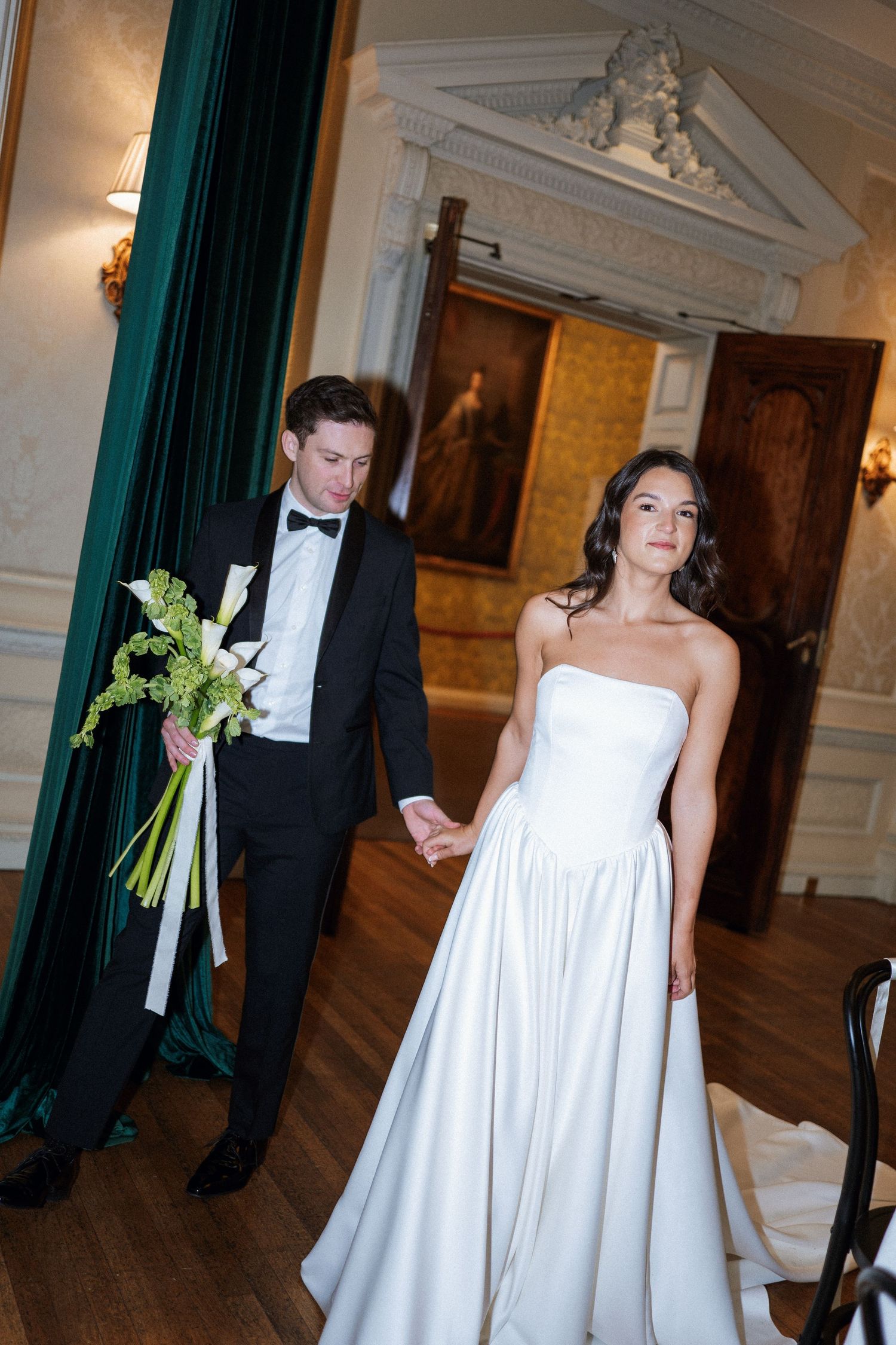 A bride and groom walk through an elegant hallway with green curtains and ornate wall decor during their wedding.