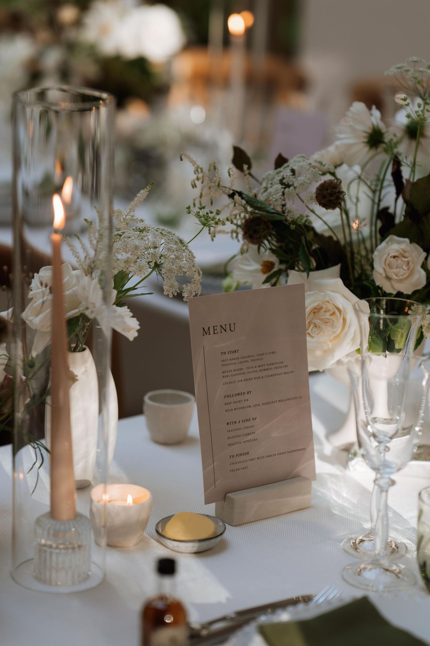 Elegant wedding table setting with menu card, white roses, candles and delicate glassware on white tablecloth.