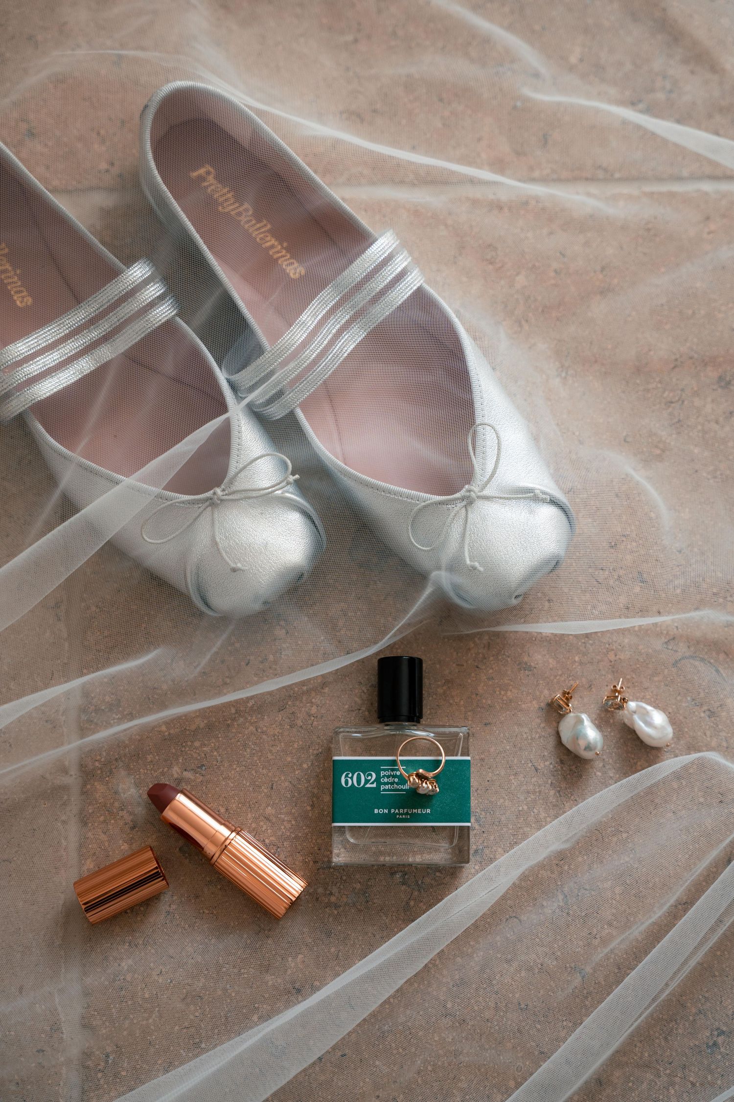 Silver ballet flats with delicate tulle ribbons and copper lipstick tubes arranged on a beige textured surface.