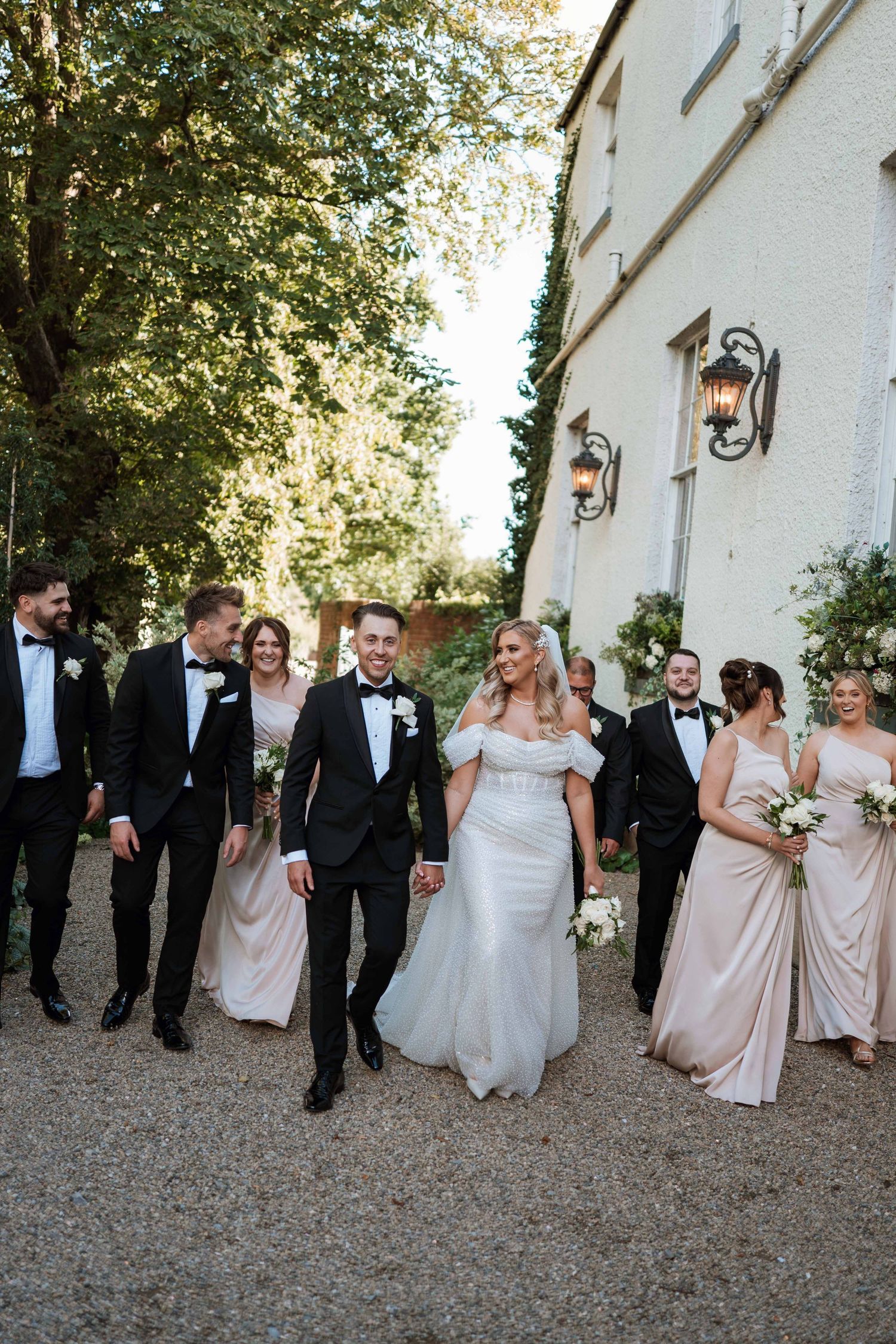 Wedding party in formal attire walking and laughing together along a gravel path beside a white building.