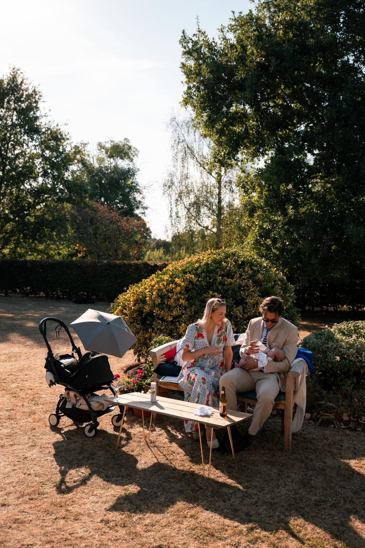 A couple enjoys a sunny outdoor picnic on a bench with a baby stroller nearby in a garden setting.