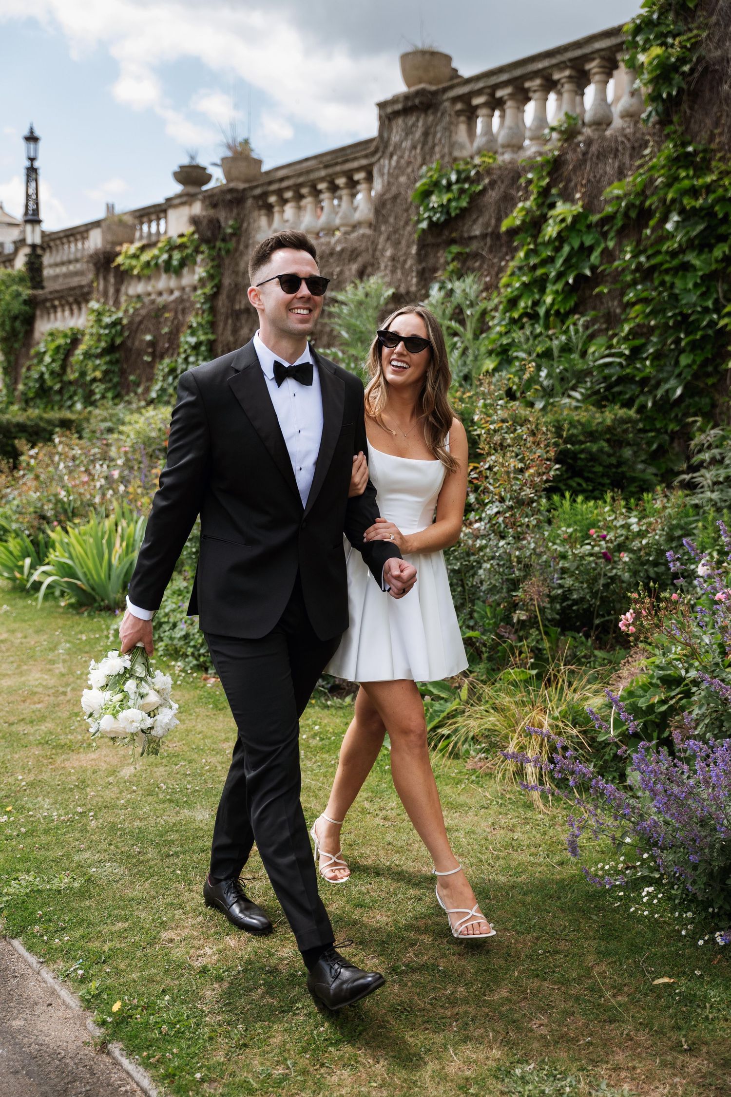 A couple in formal attire walking together in a garden with stone balustrades and flowering plants.