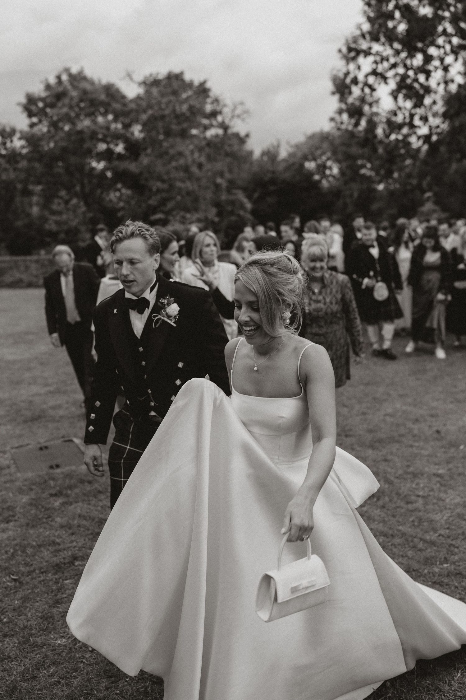 A black and white photo shows wedding guests gathering outdoors while a bride in a flowing white dress walks past.