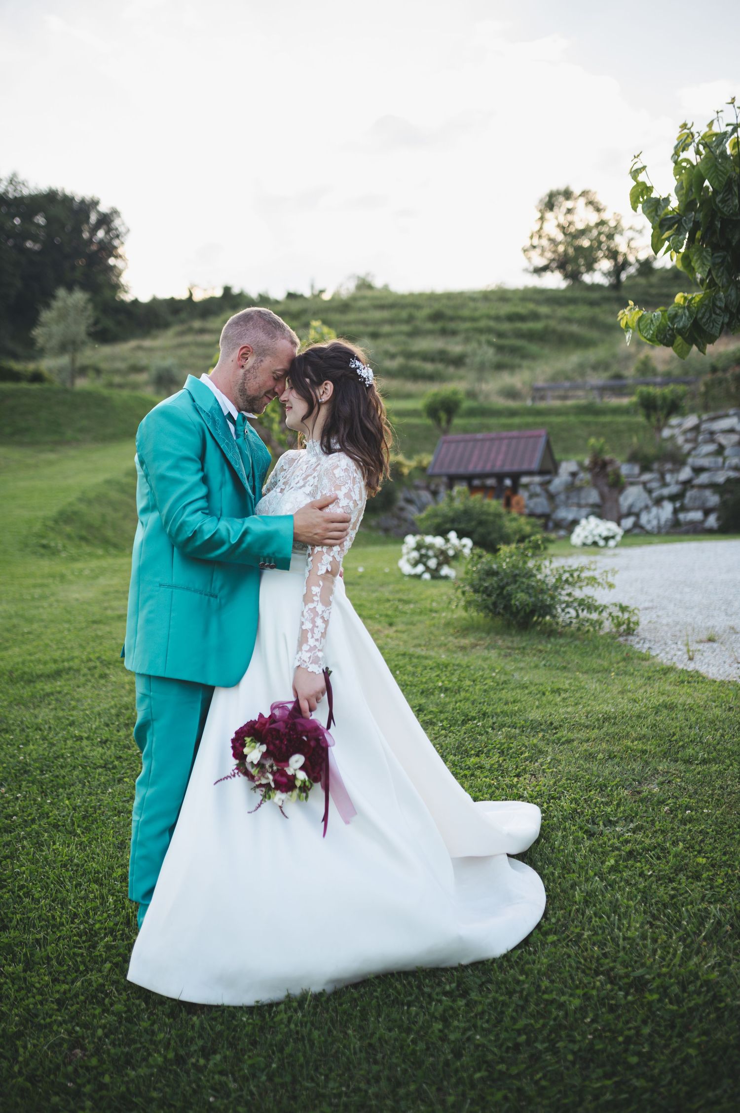 Couple in turquoise suit and white lace wedding dress embrace on grassy hill during outdoor wedding photo session.