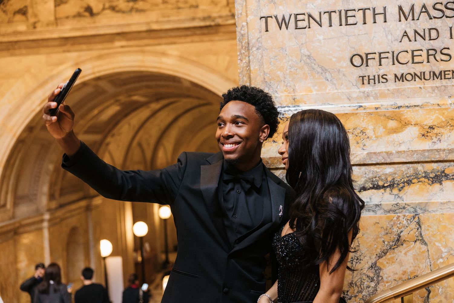 A young couple in formal black attire takes a selfie inside a grand historic building with ornate architecture.