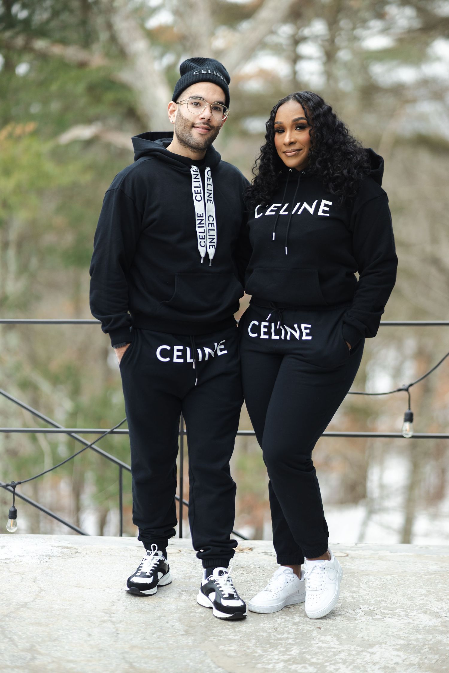 Couple poses in matching black Celine hoodies and sneakers against a winter forest backdrop.