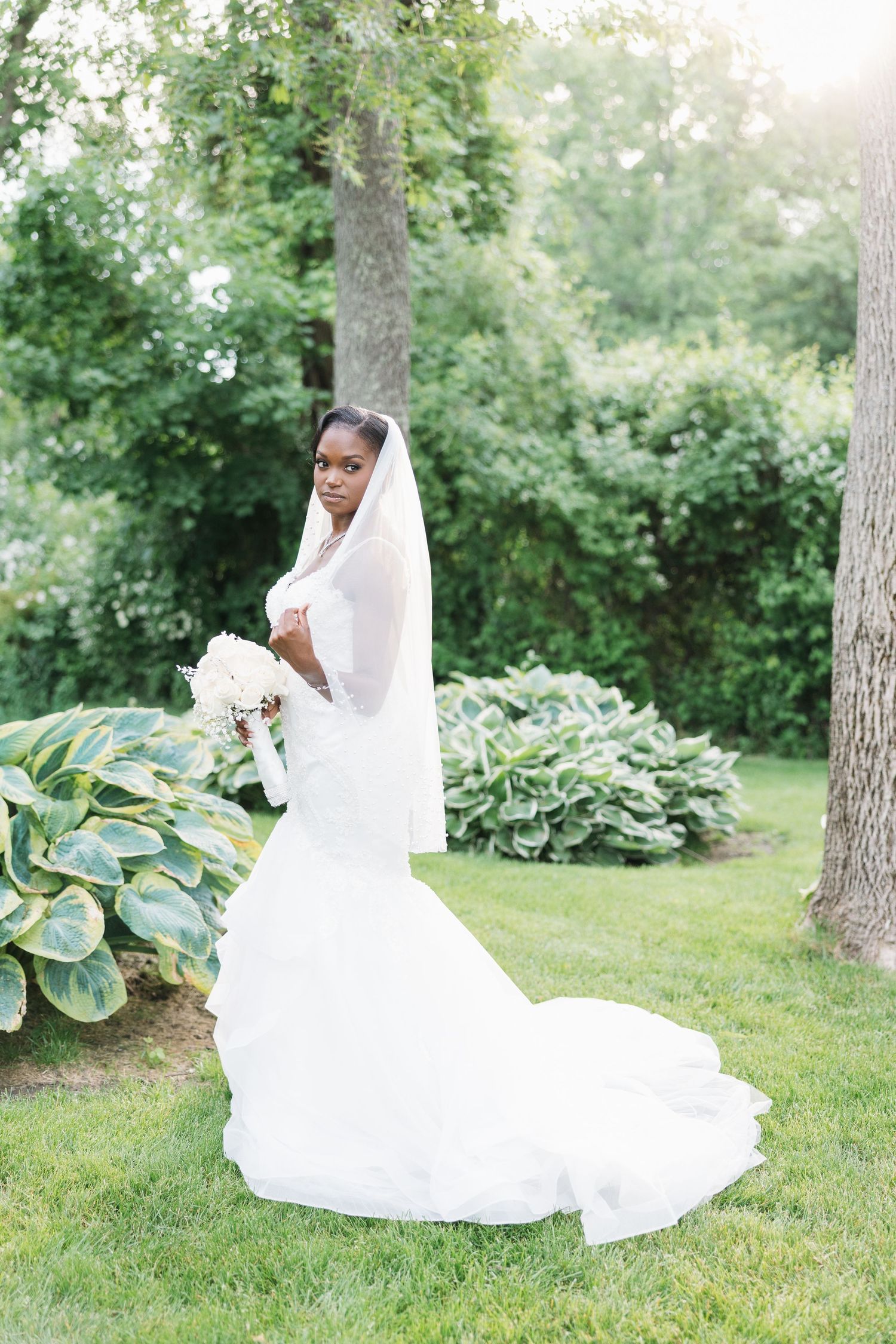 Elegant bride in white mermaid gown and veil holds bouquet in lush garden setting.