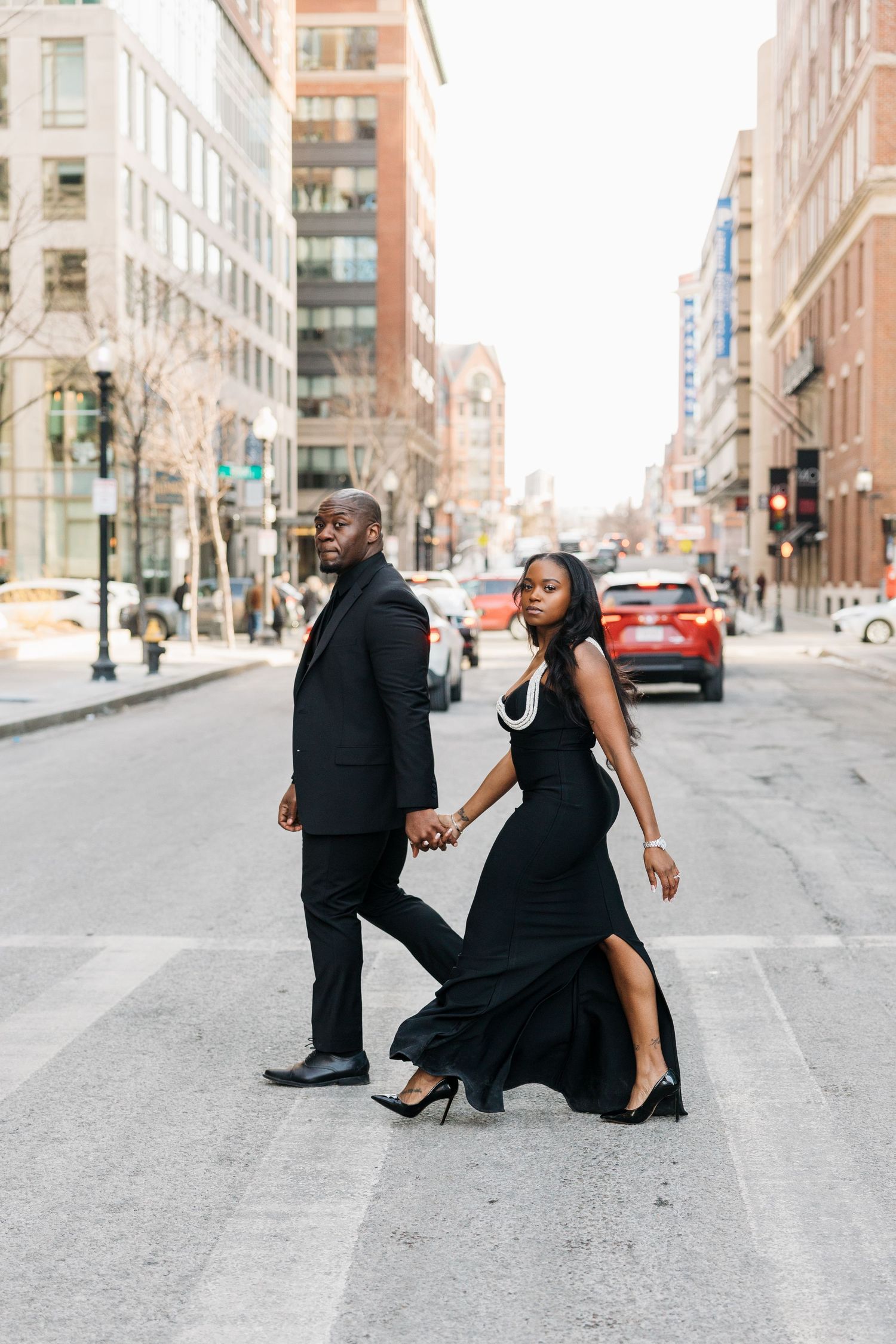 Elegant couple in matching black formal attire crossing a city street, holding hands in an urban setting.