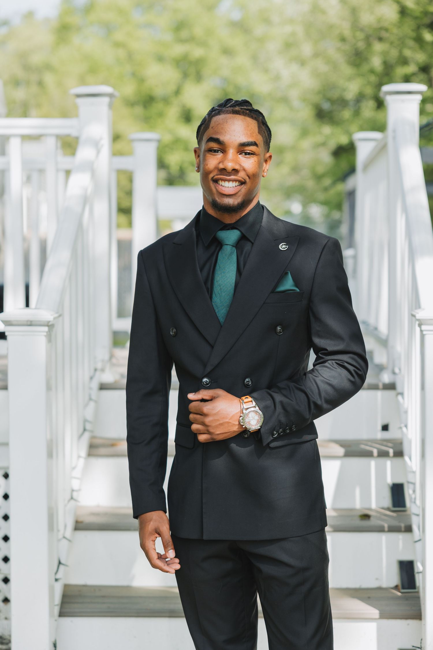 A person in a black formal suit and emerald green tie poses confidently against a backdrop of white pillars outdoors.