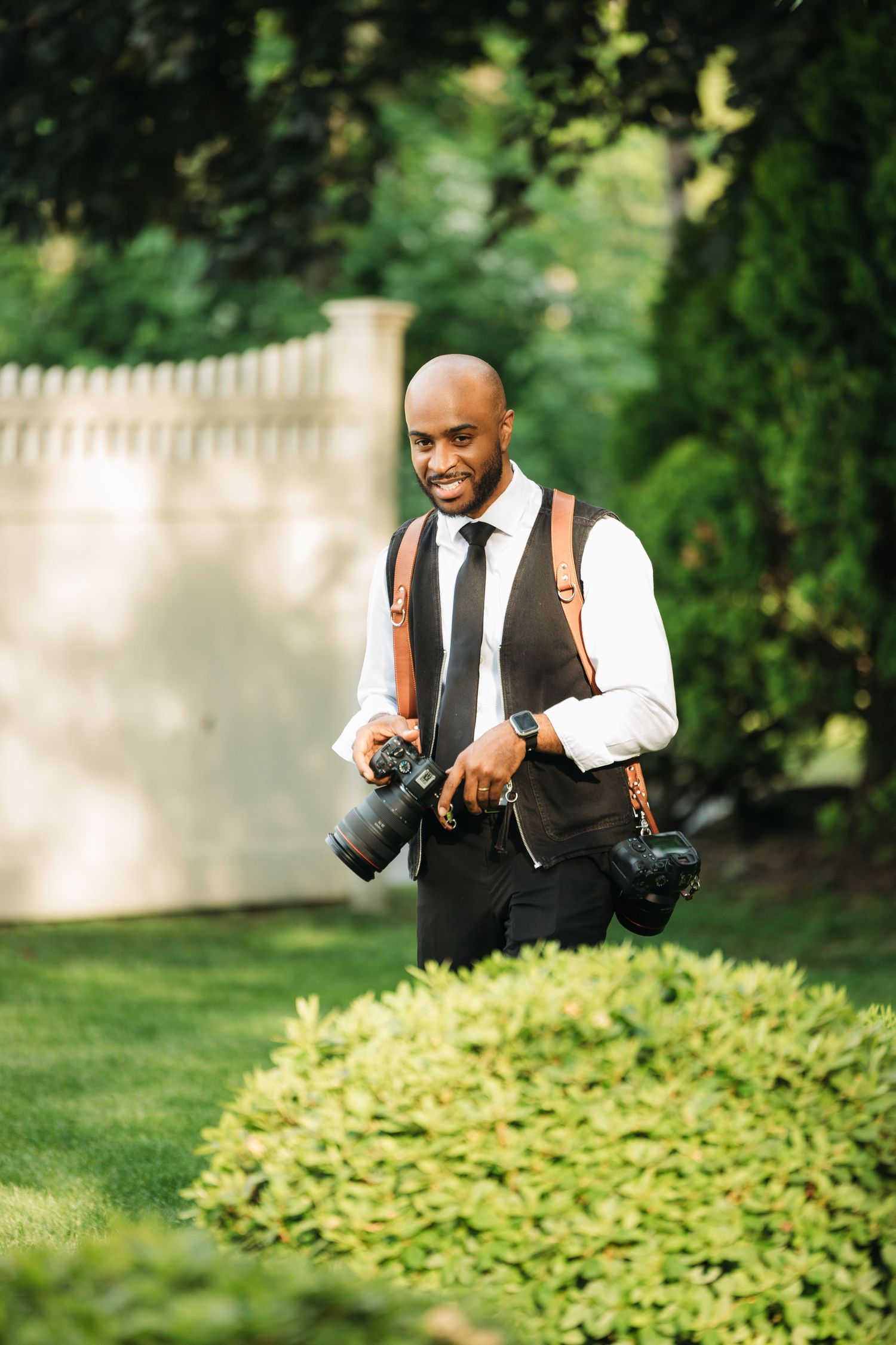 Smiling bald Black male photographer holding DSLR cameras with leather straps in a lush green garden.