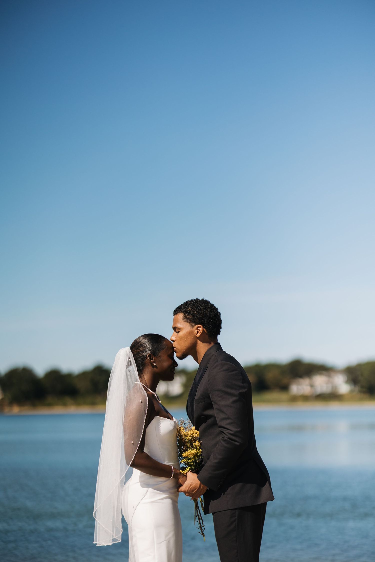 A couple shares a romantic kiss by a serene waterfront on their wedding day with a bright blue sky in the background.