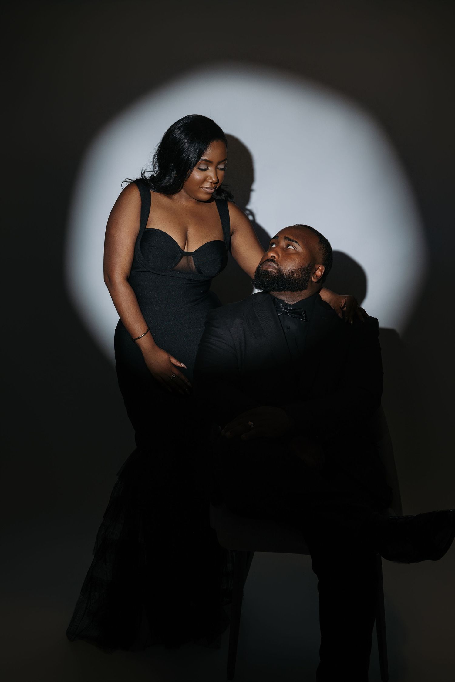Stylish couple in black formal wear pose intimately against dramatic spotlight in dark studio setting.