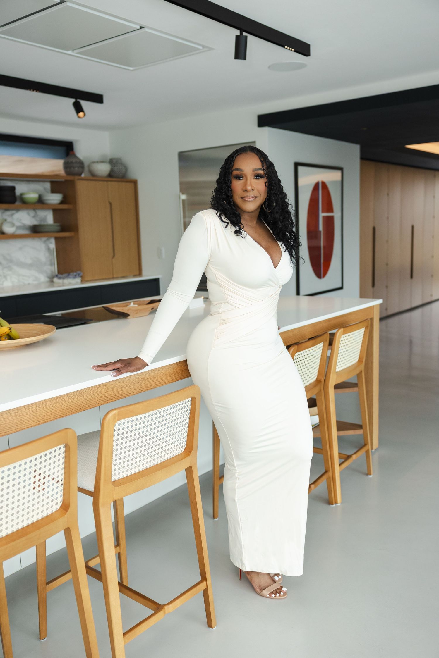 Person in a sleek white long-sleeved dress poses elegantly at a modern kitchen counter with wooden bar chairs.