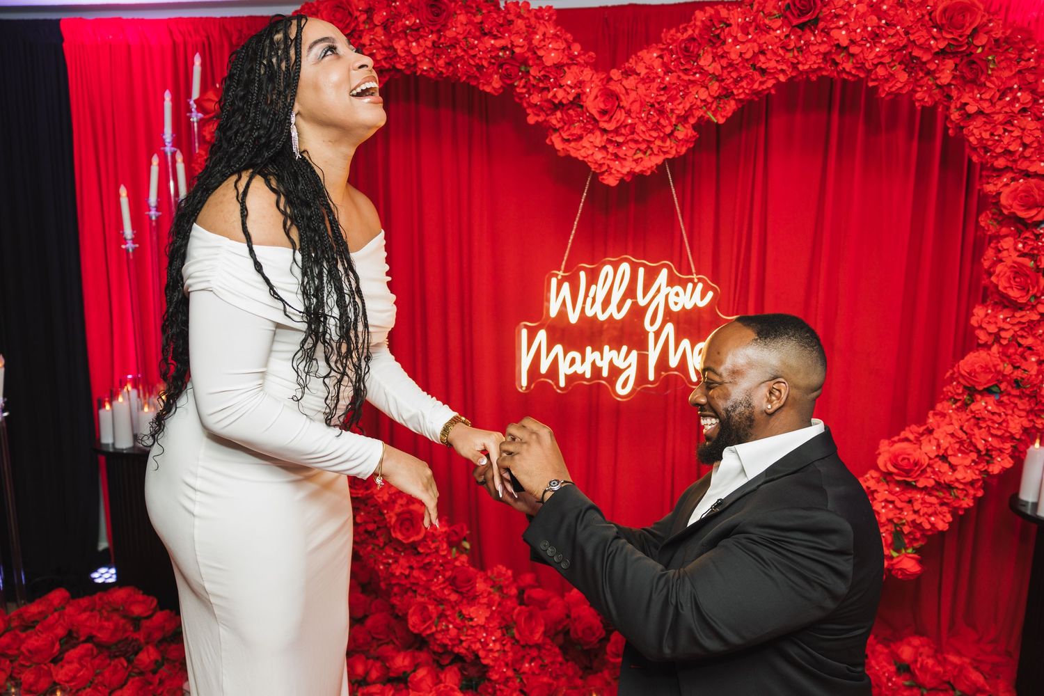 A romantic marriage proposal scene with a red heart backdrop and illuminated 'Will You Marry Me' sign glowing on the wall.