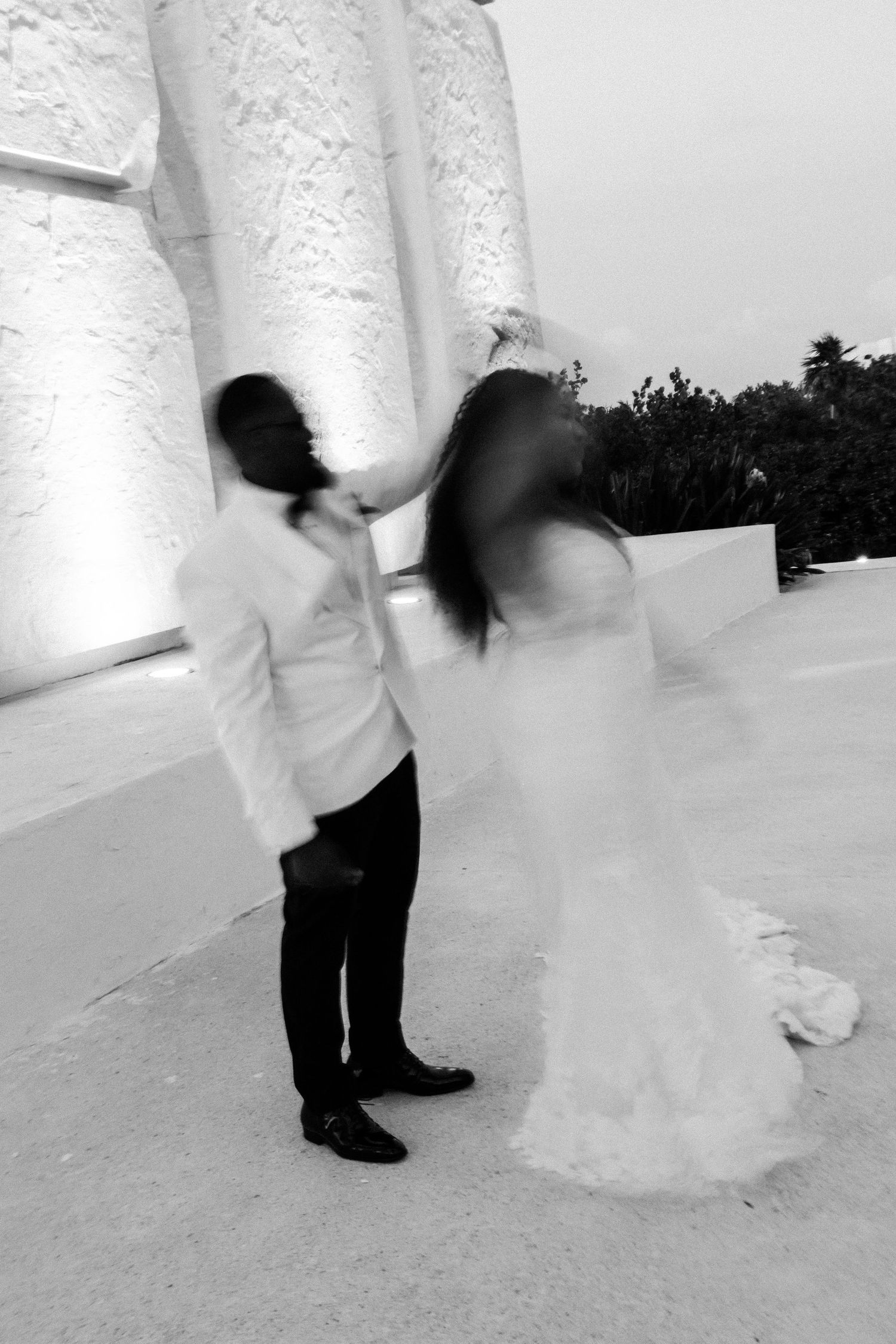 Black and white photo of a bride and groom dancing outdoors at their wedding reception near illuminated columns.