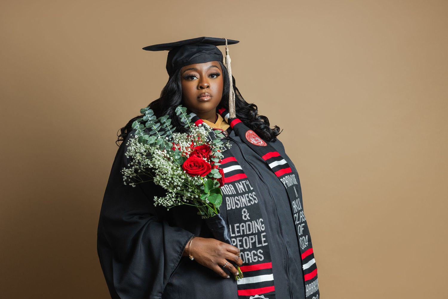 Graduate in black cap and gown holds red and white bouquet while posing for professional graduation photos.