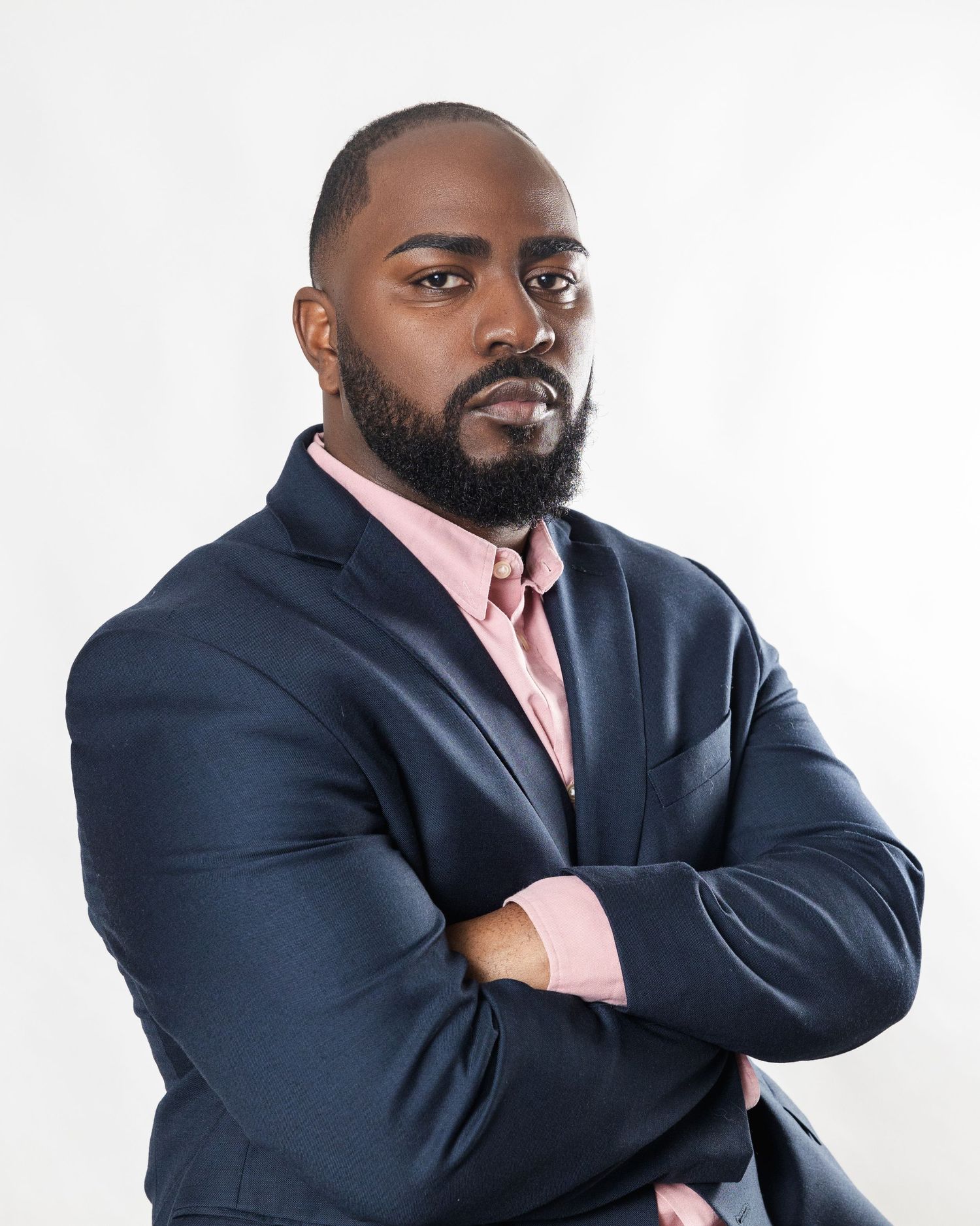 Professional headshot of someone in a navy suit and pink dress shirt posing with arms crossed against white background.