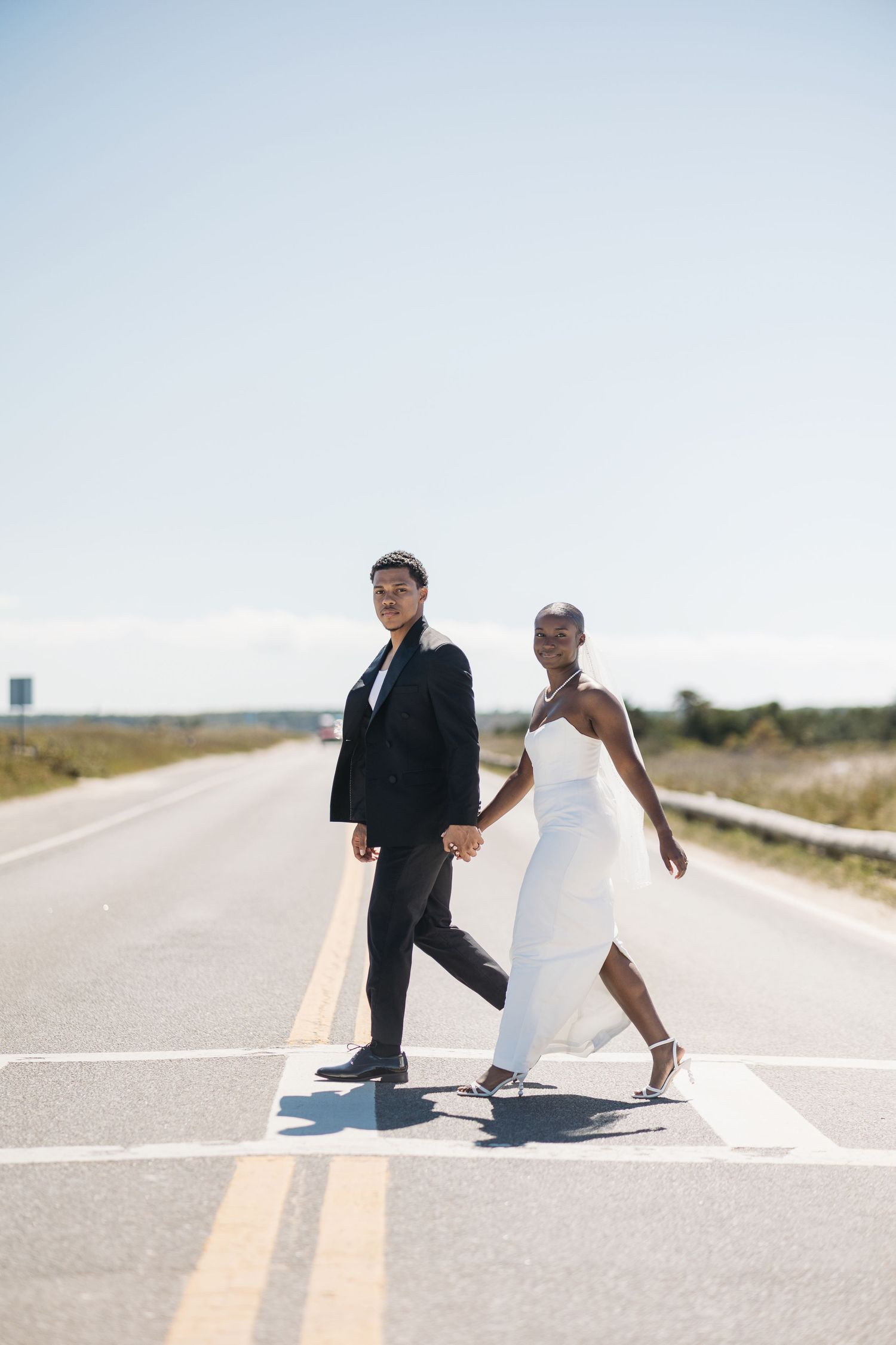 A couple in wedding attire holds hands while crossing a road, the bride in white and groom in a black suit.
