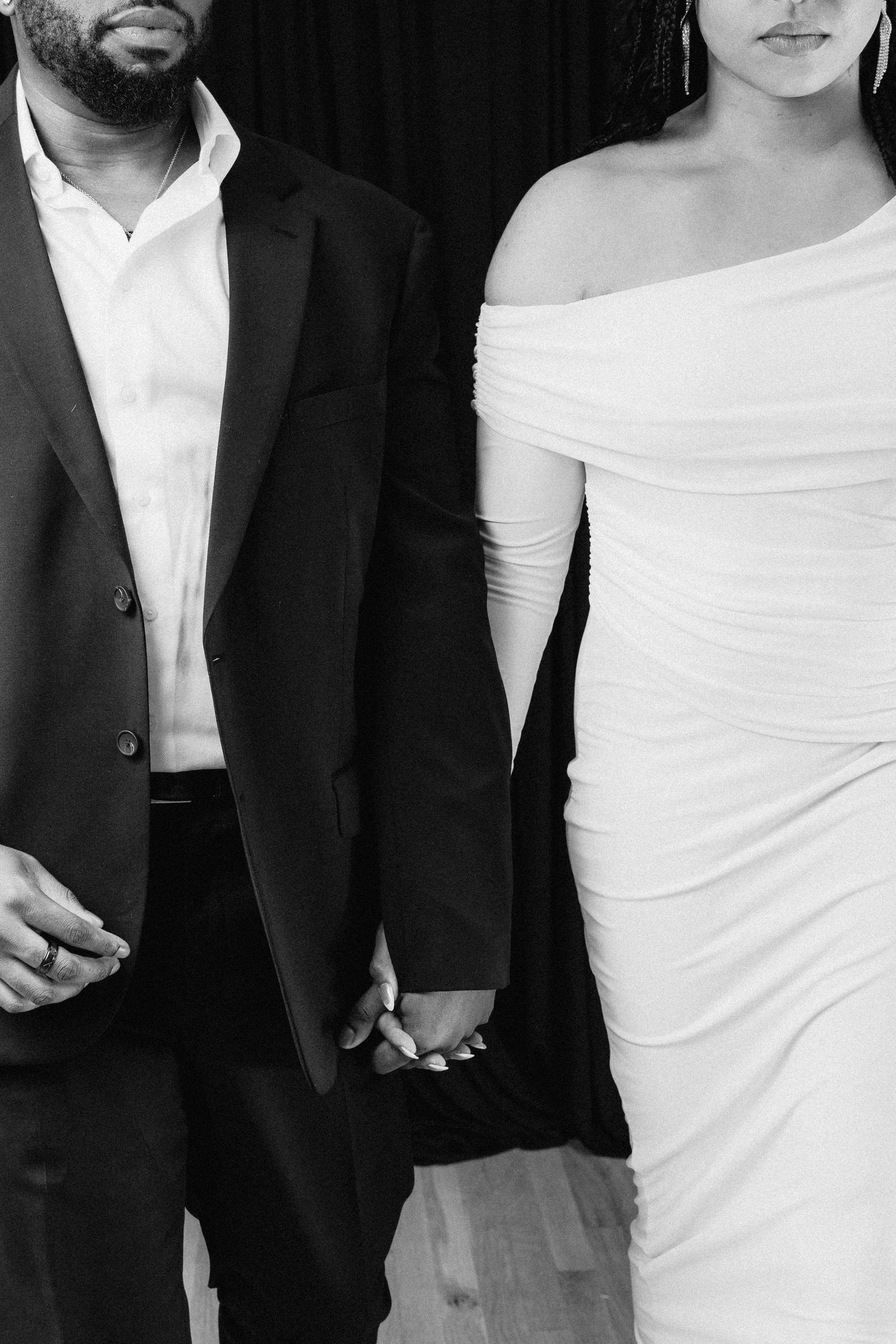 Black and white photo showing couple holding hands, with person in dark suit and person in white off-shoulder dress.
