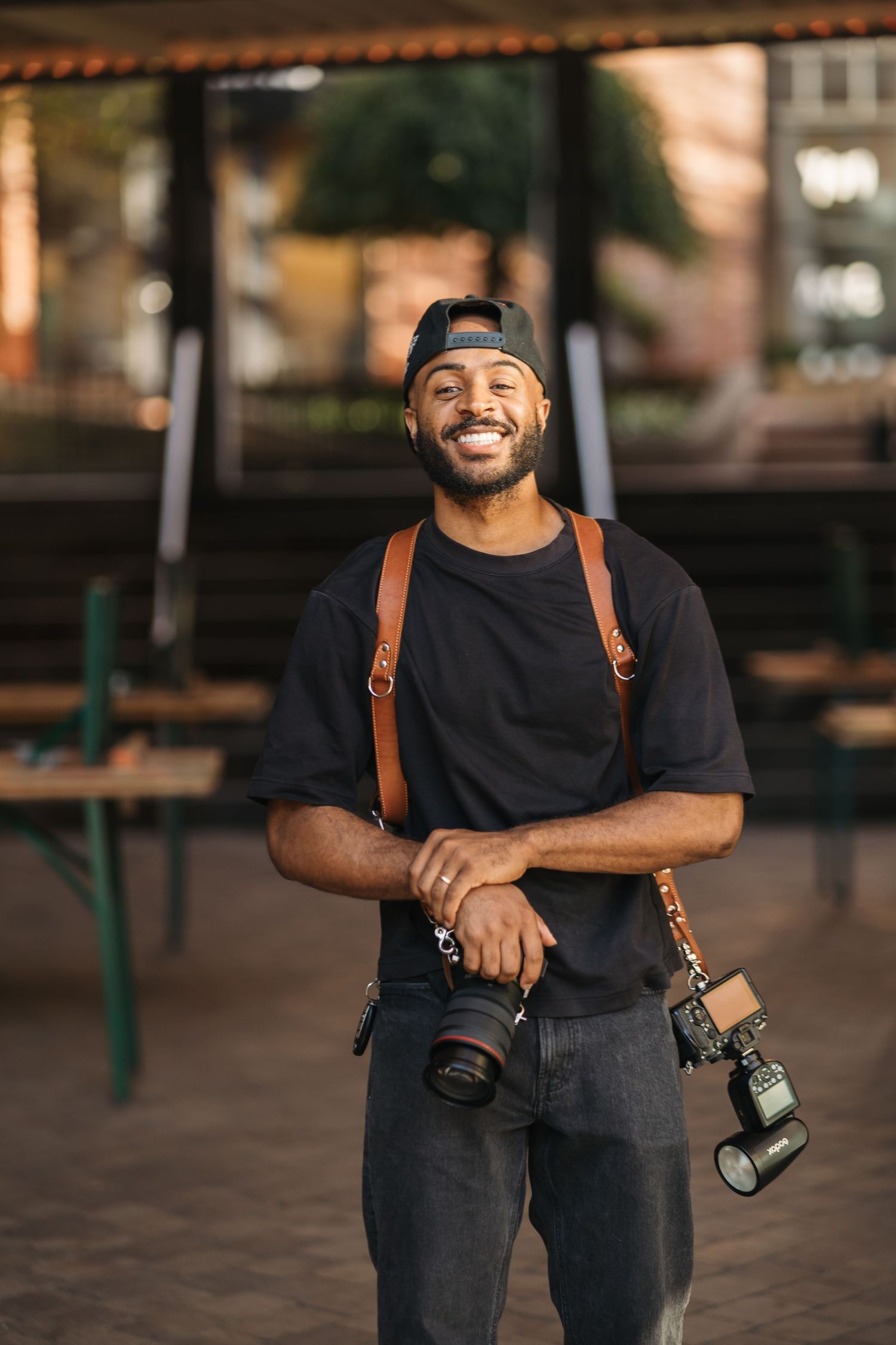 Smiling Black photographer holding DSLR camera with brown leather strap, wearing black t-shirt and cap outdoors.