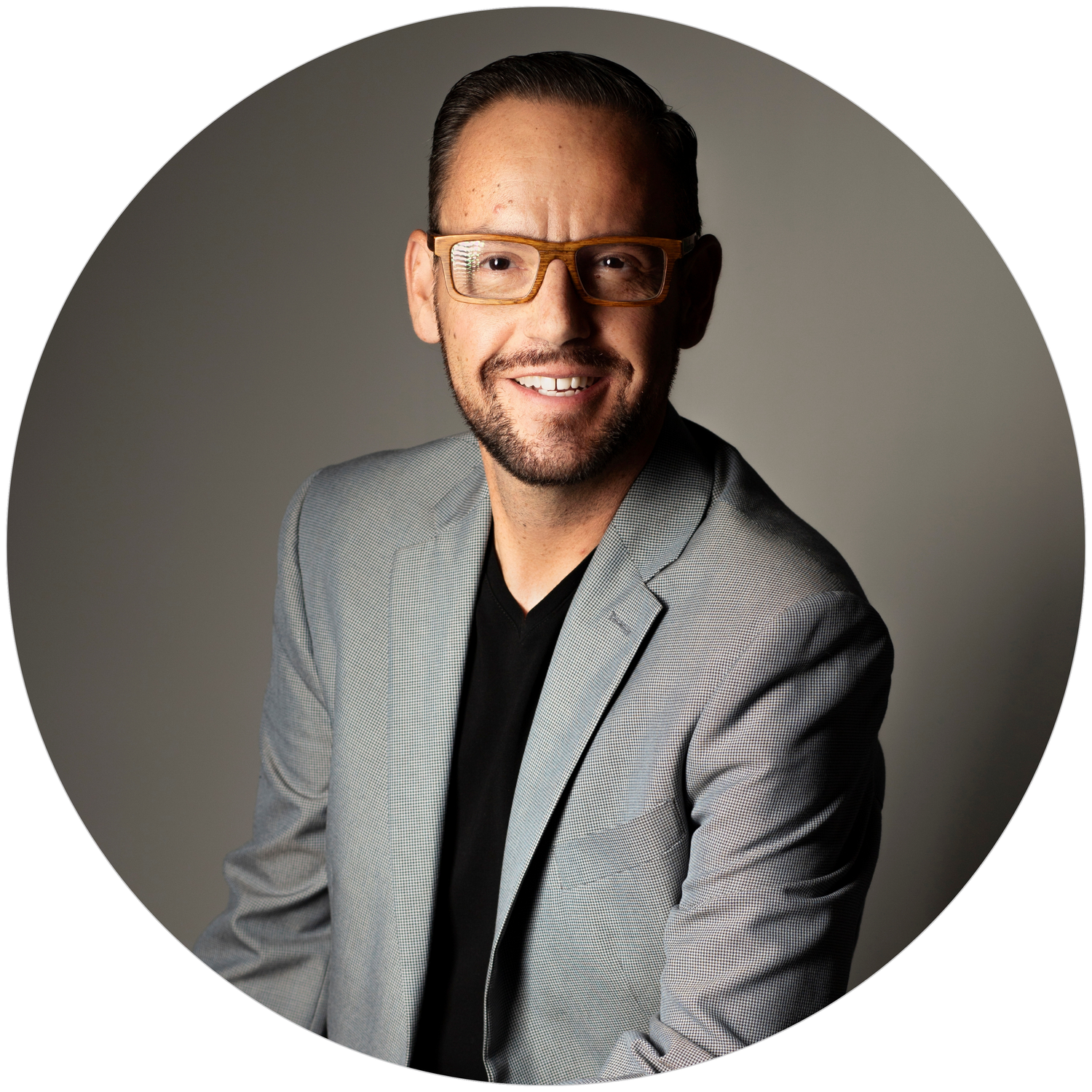 Professional man in gray blazer and glasses smiling confidently against neutral background.