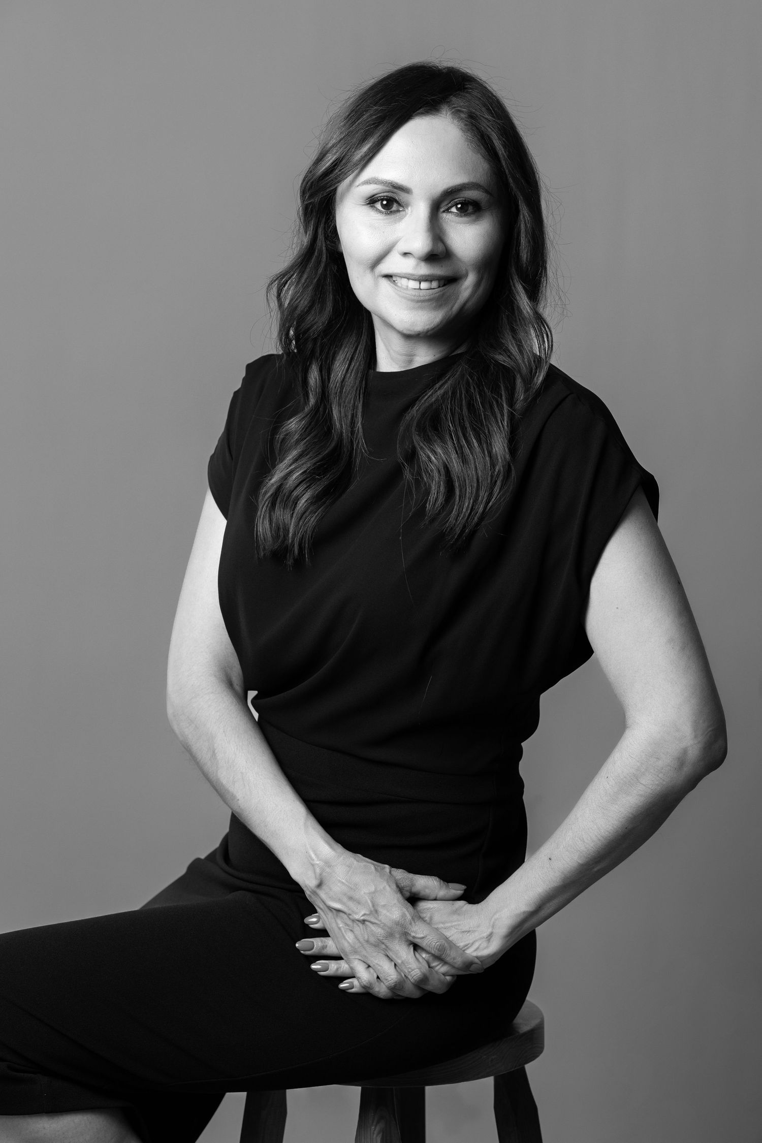 Black and white portrait of a smiling woman in black top seated on stool with hands clasped.