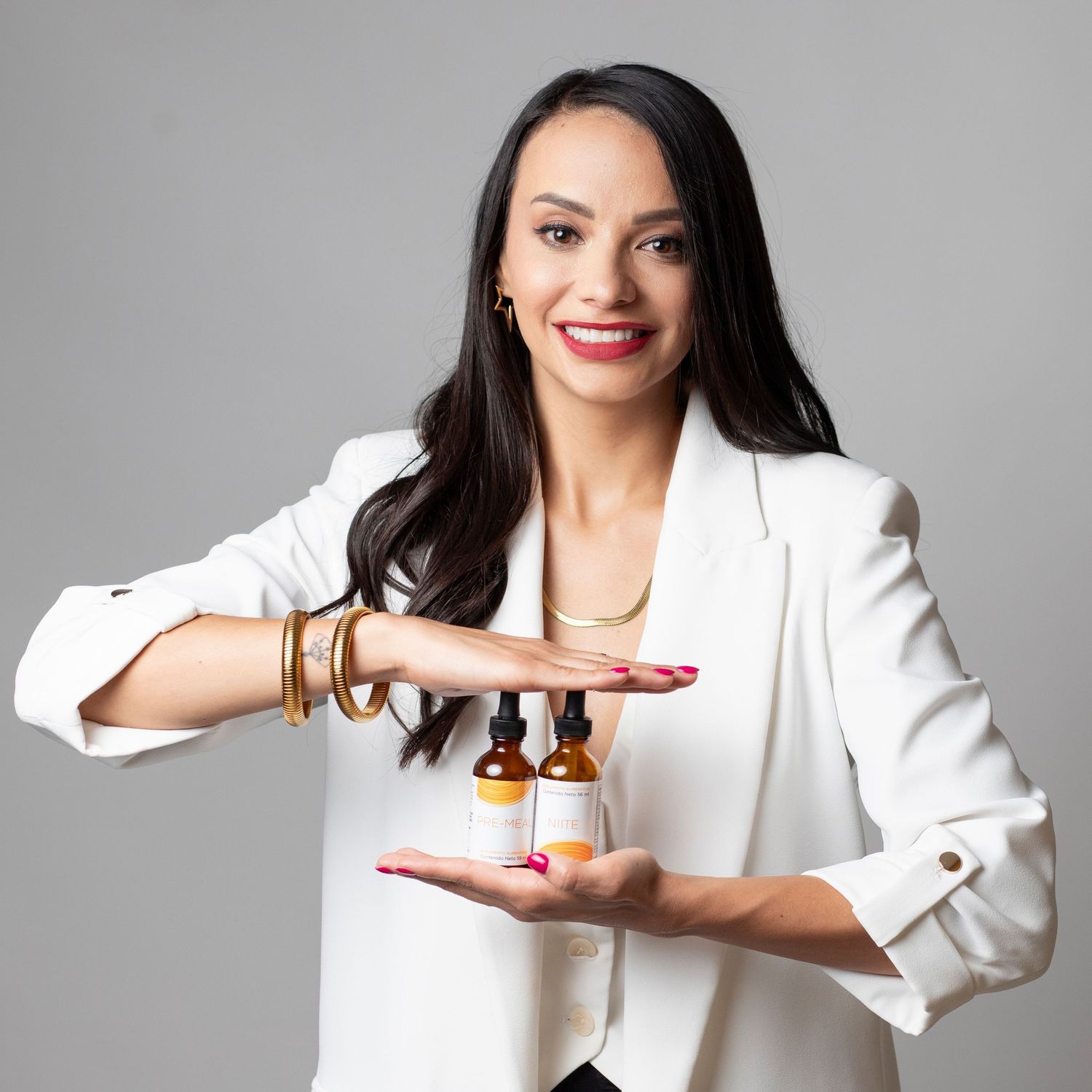 Professional in white blazer displays essential oil bottles with confident smile against neutral background.