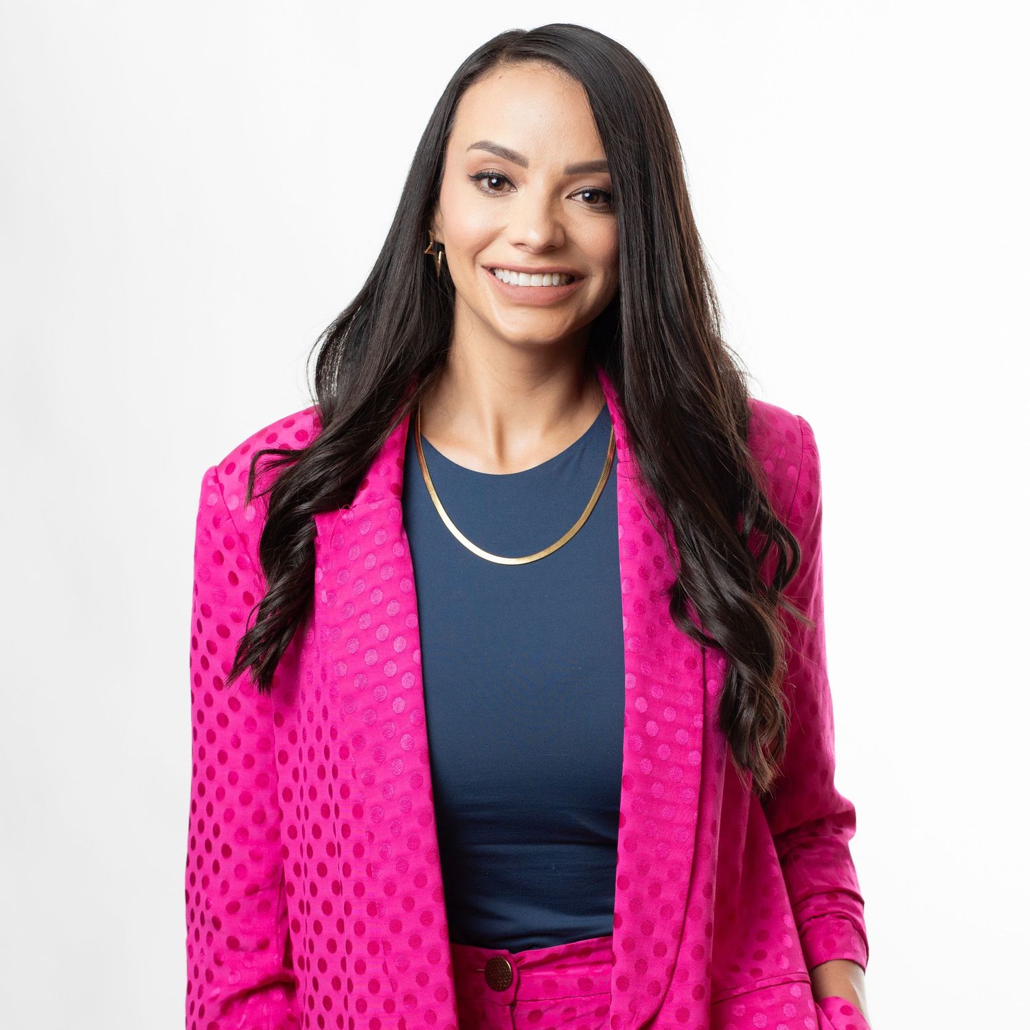 Professional woman in bright pink blazer with polka dot pattern and navy top smiles against white background.