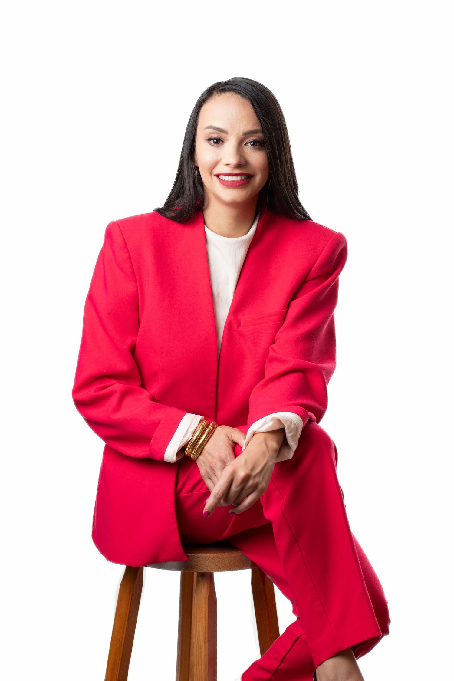 Professional woman in vibrant red blazer sitting confidently on wooden stool against white background.