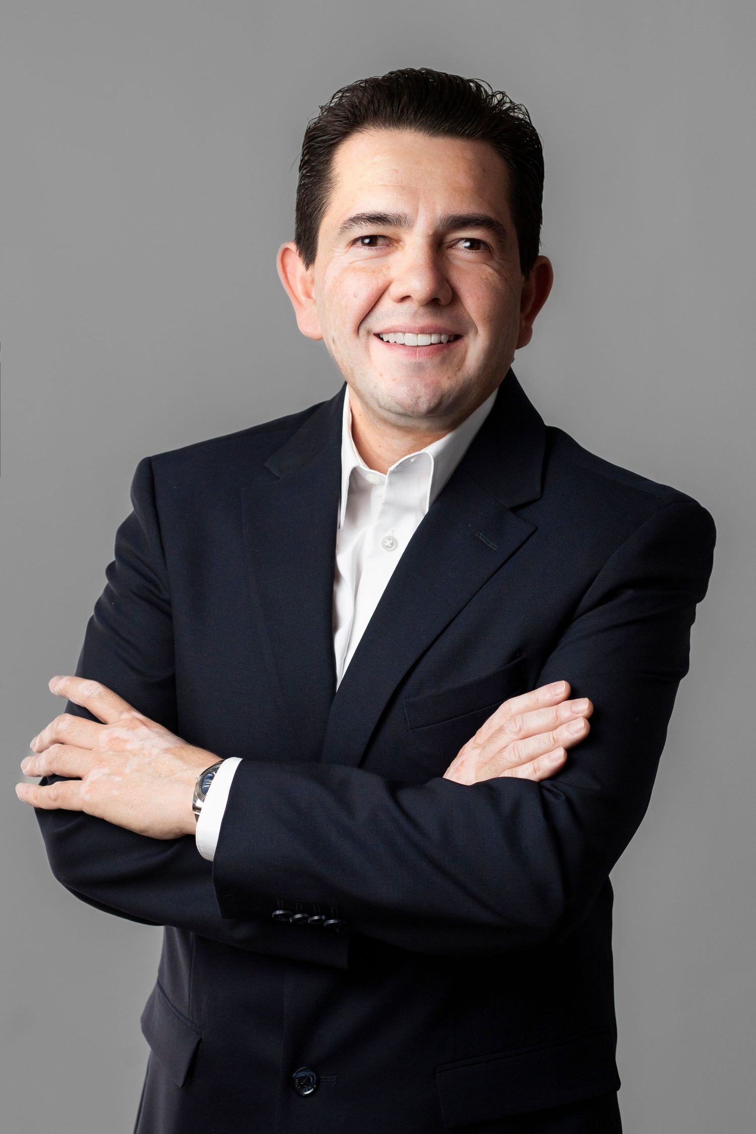 Professional headshot of someone in a dark suit and white shirt with arms crossed smiling against gray background.