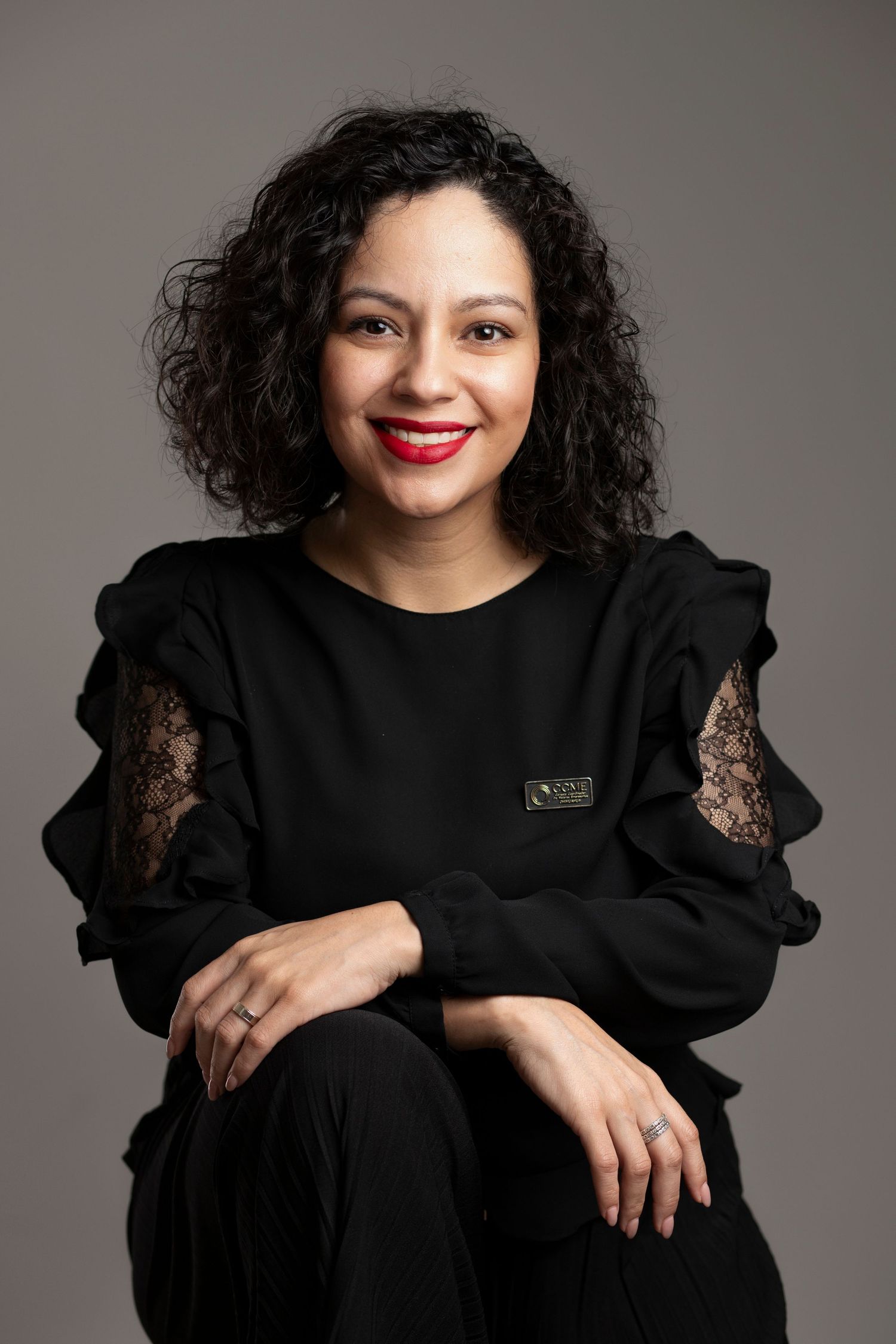 Woman in black ruffled lace top smiling confidently, seated against gray background, wearing red lipstick.