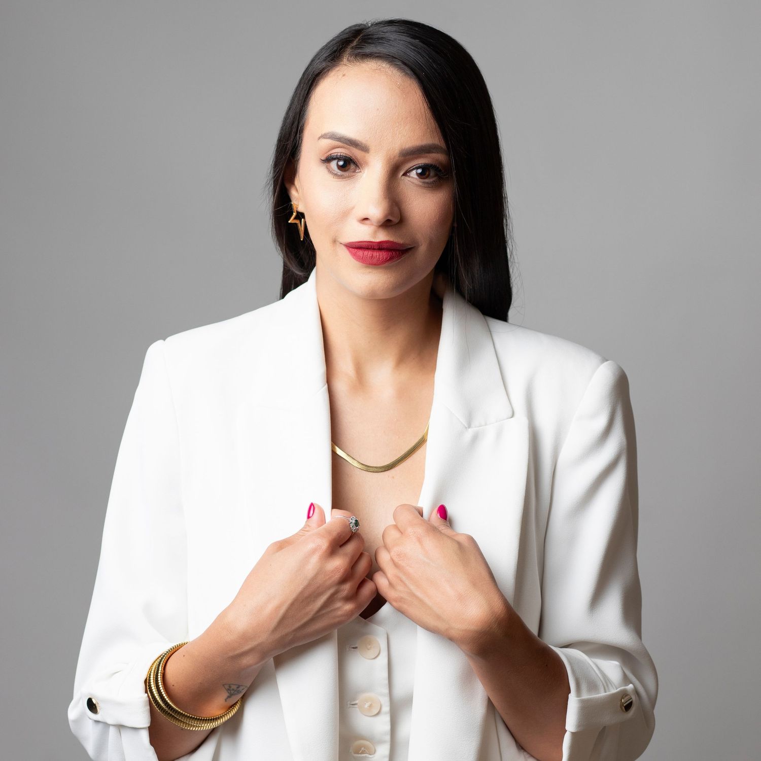 Professional portrait of someone in a crisp white blazer and gold jewelry against a gray background.