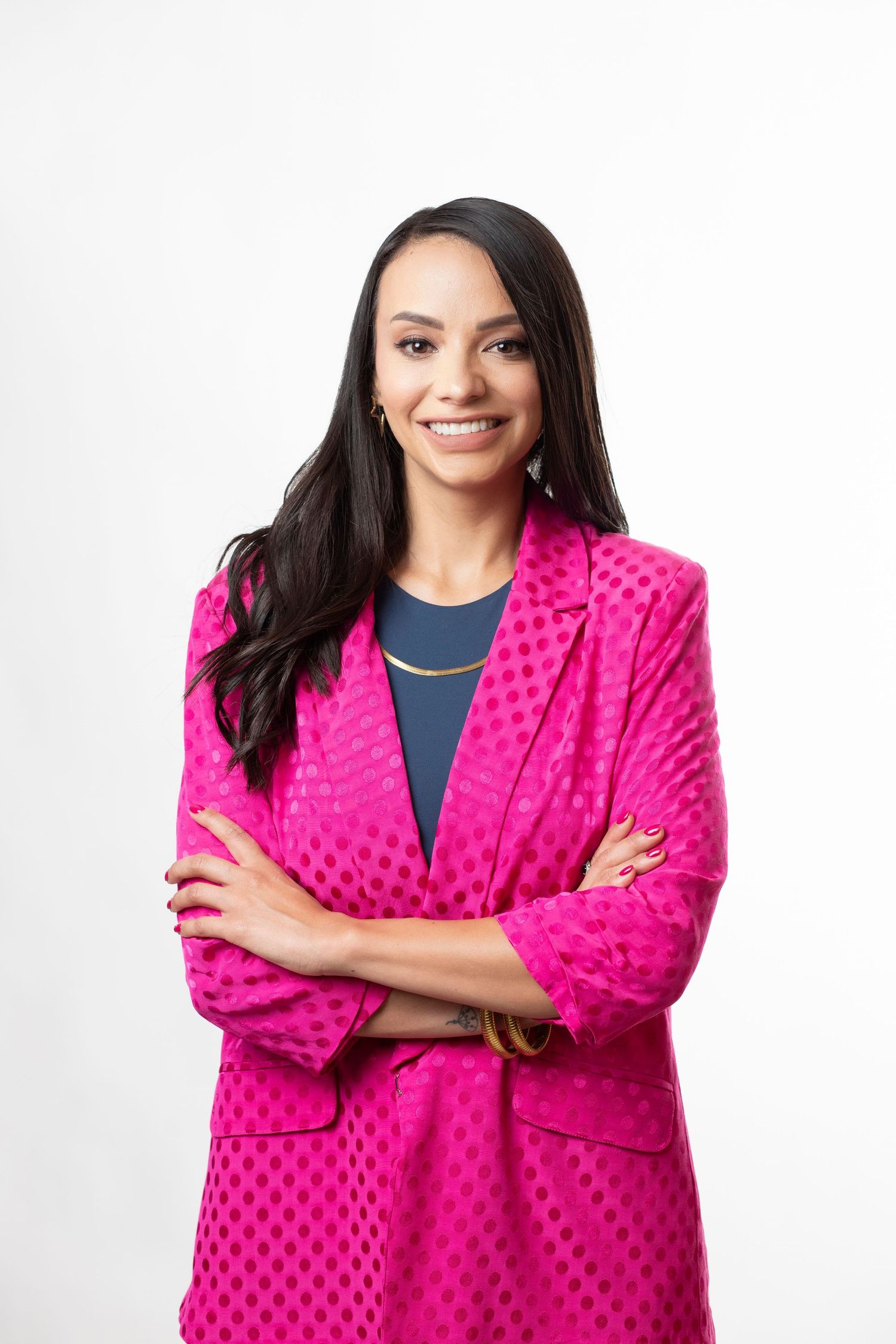 Woman wearing bright pink polka dot blazer poses confidently with arms crossed against white background.