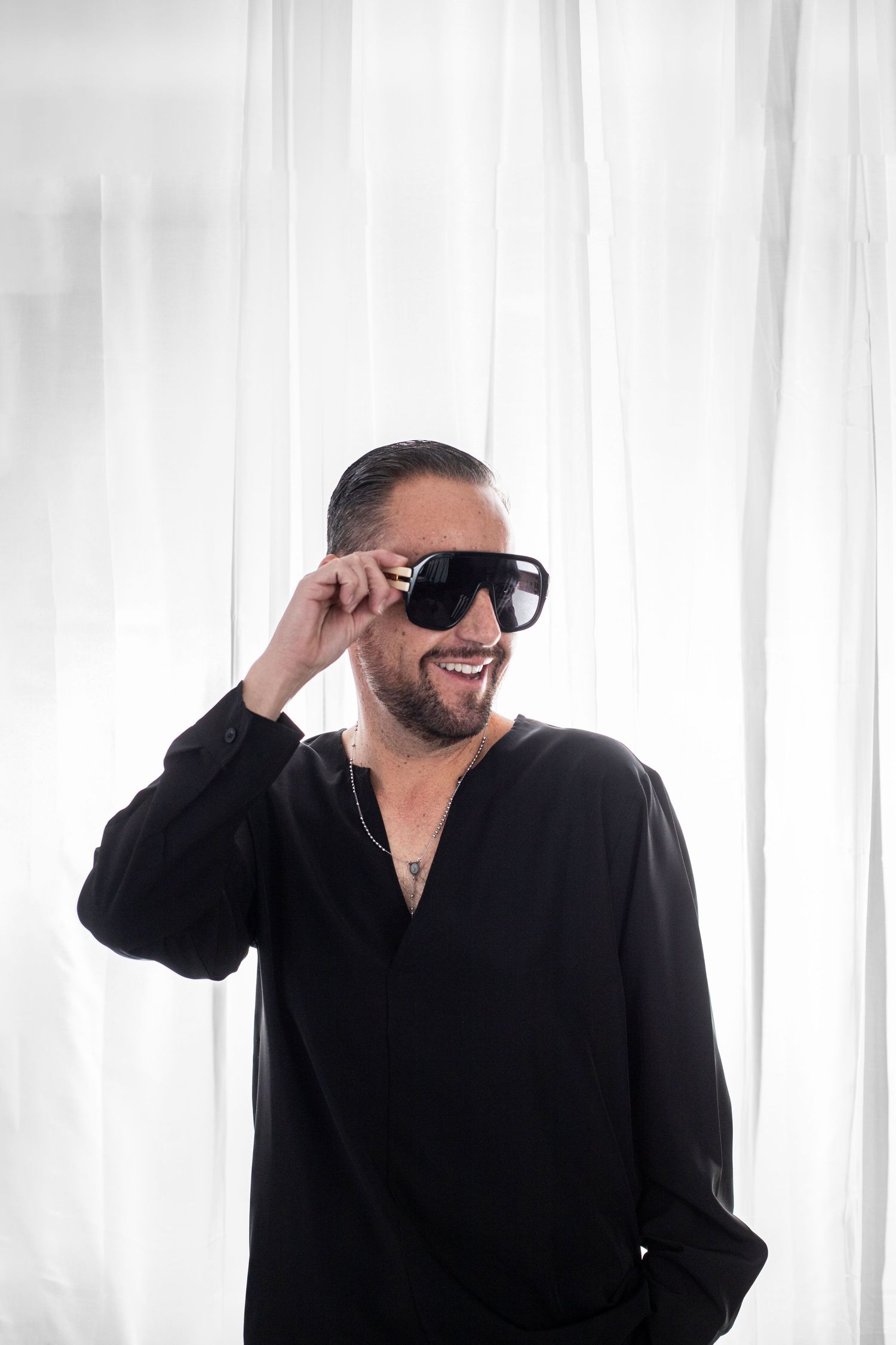 Person wearing black shirt and sunglasses smiles while adjusting eyewear against white curtain background.