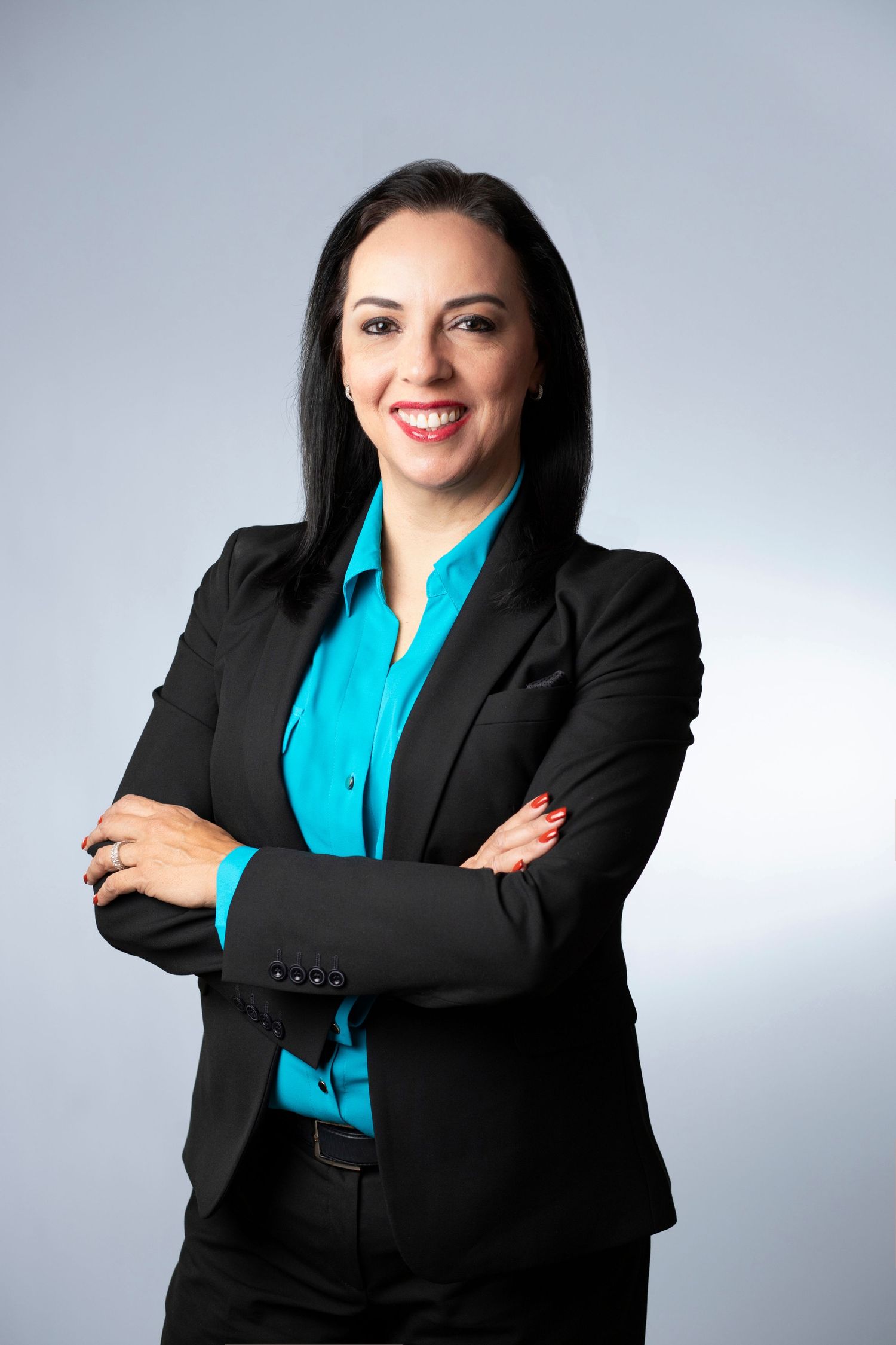 Professional business portrait of someone in a black blazer and turquoise blouse standing with arms crossed against gray backdrop.