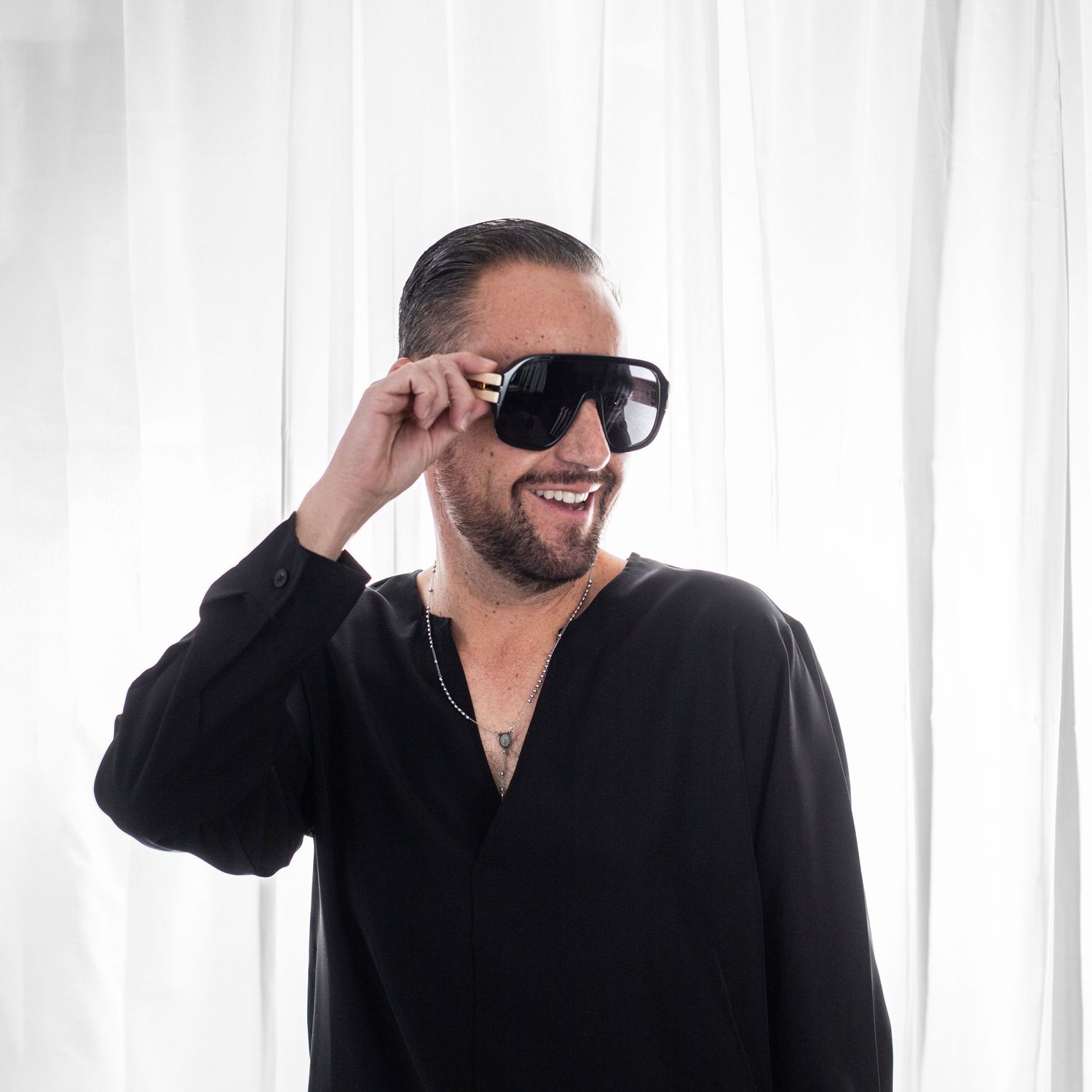 Smiling person wearing dark sunglasses and black shirt against white background.