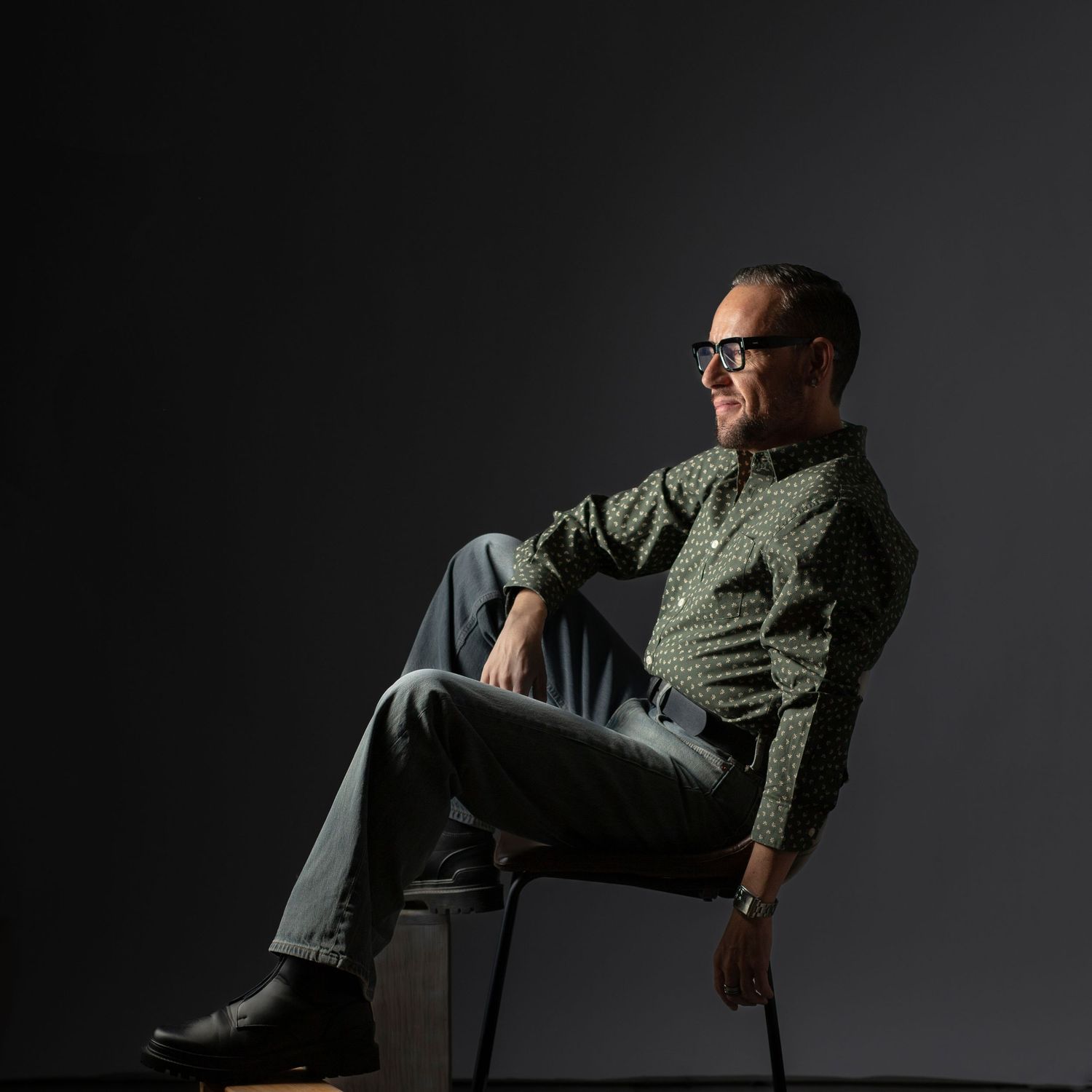 Person in glasses and olive shirt sits thoughtfully in chair against dark background in dramatic lighting.