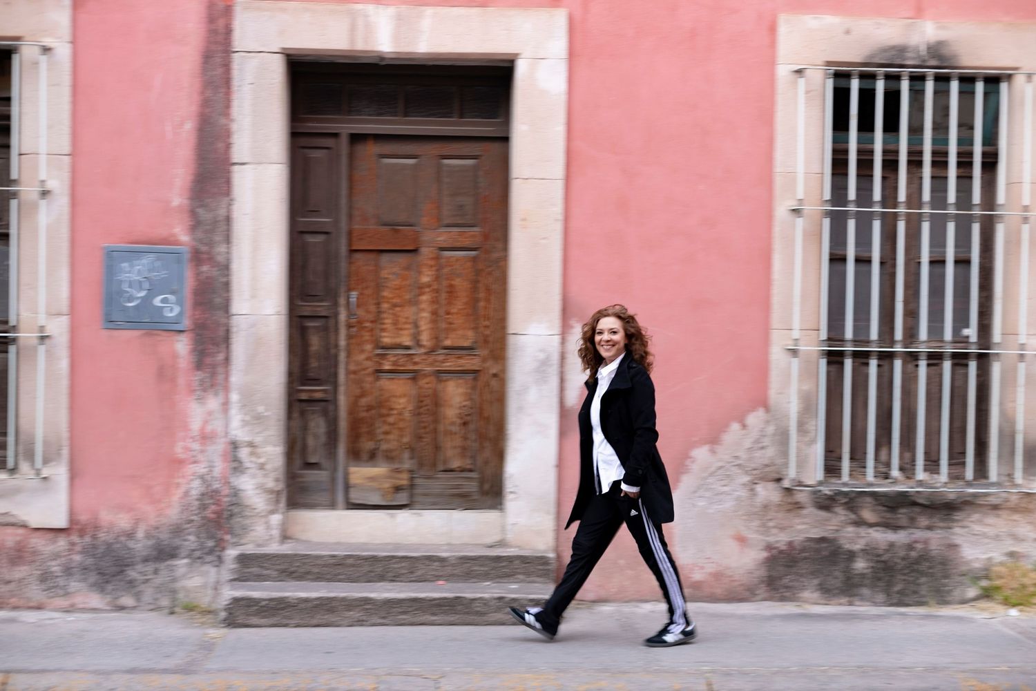 A woman in black outfit walks past a rustic pink building with wooden door and barred windows on a cobblestone street.