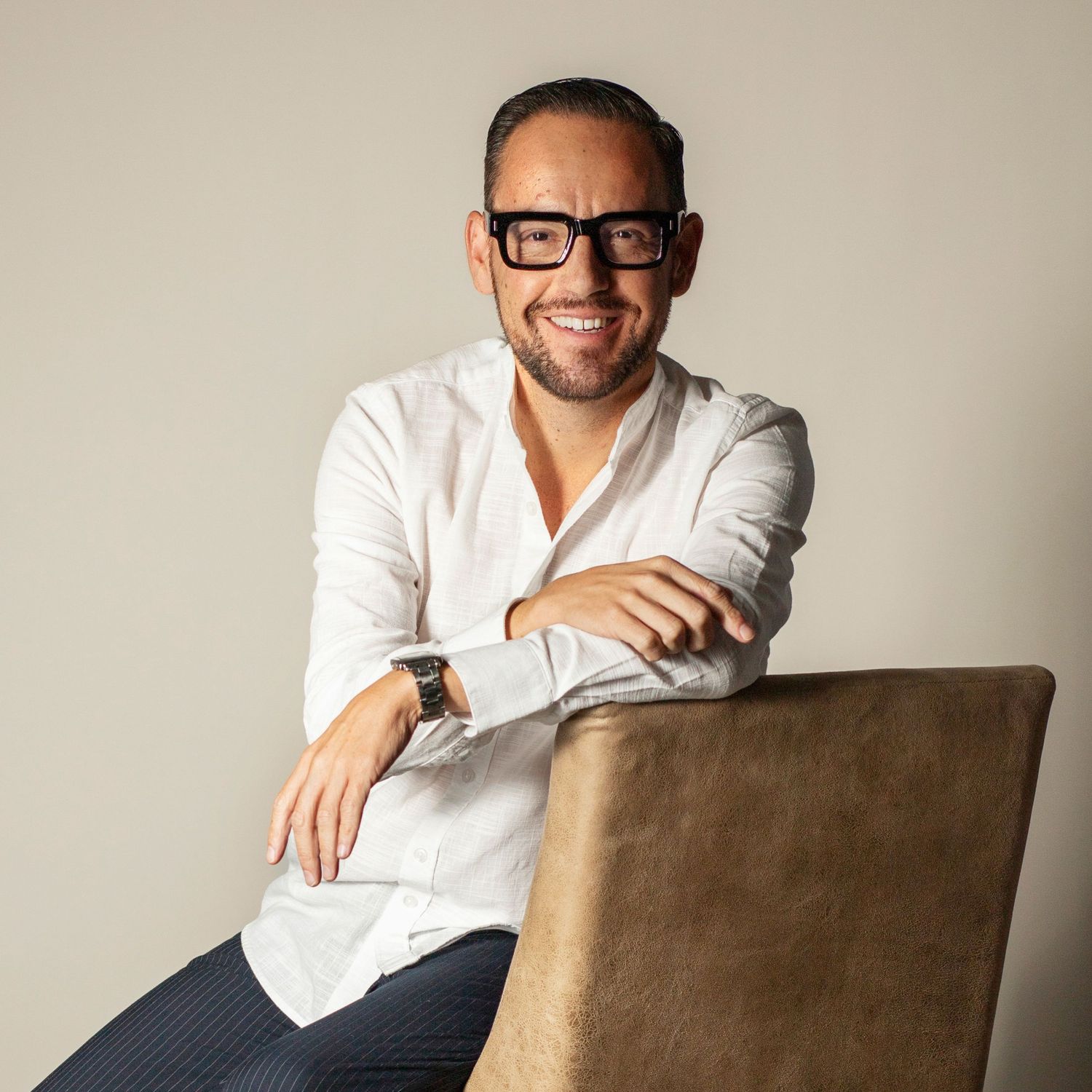 Person in white shirt and glasses sitting on brown chair smiling while posing for multiple casual portrait shots.