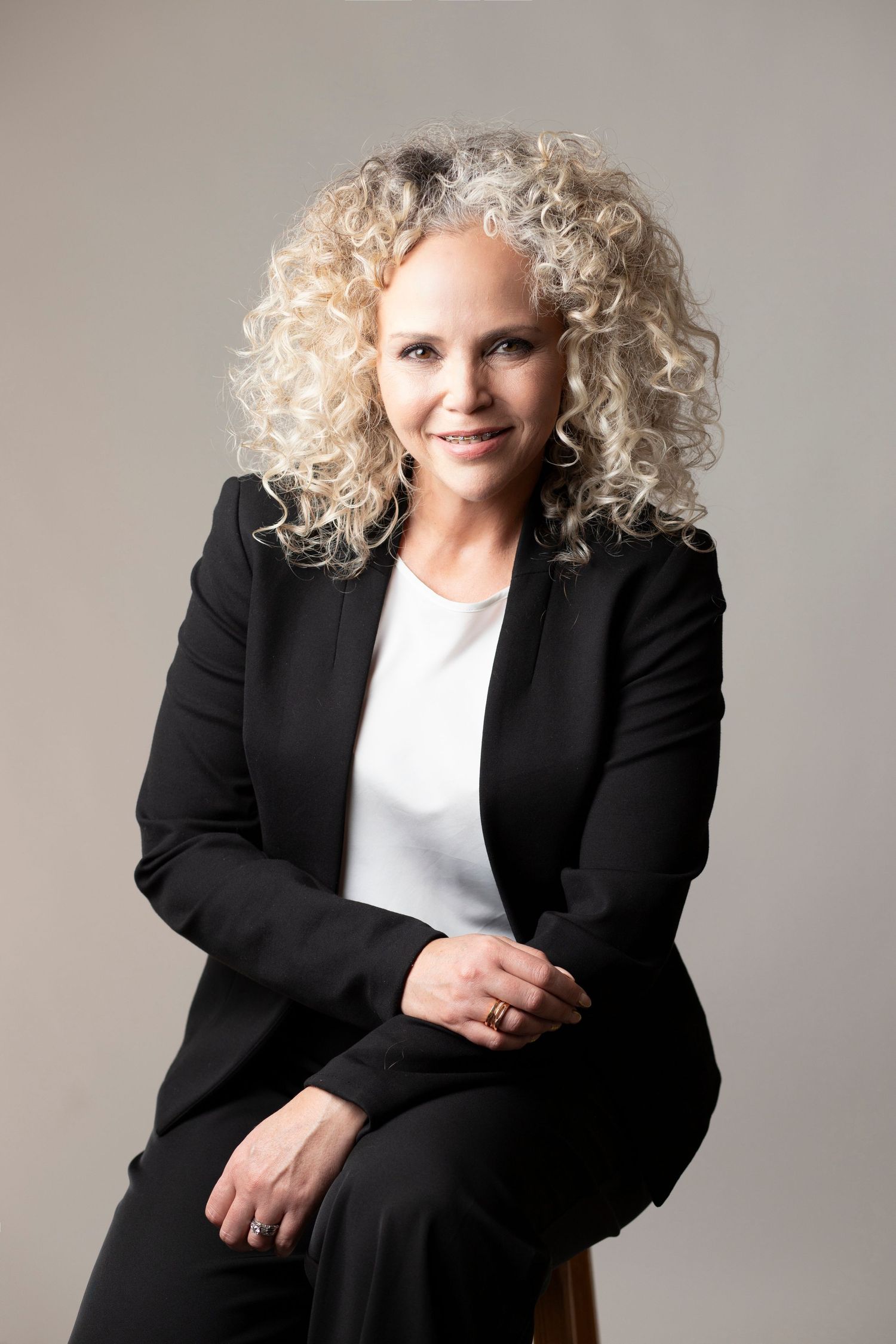 Professional headshot of a person with voluminous curly blonde hair wearing a black blazer and white top.