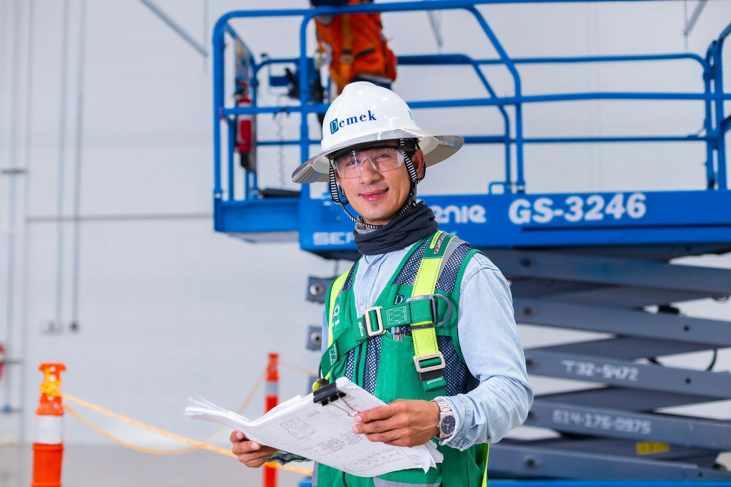 Construction worker in hard hat and safety vest reviews blueprints at a job site with aerial lift in background.