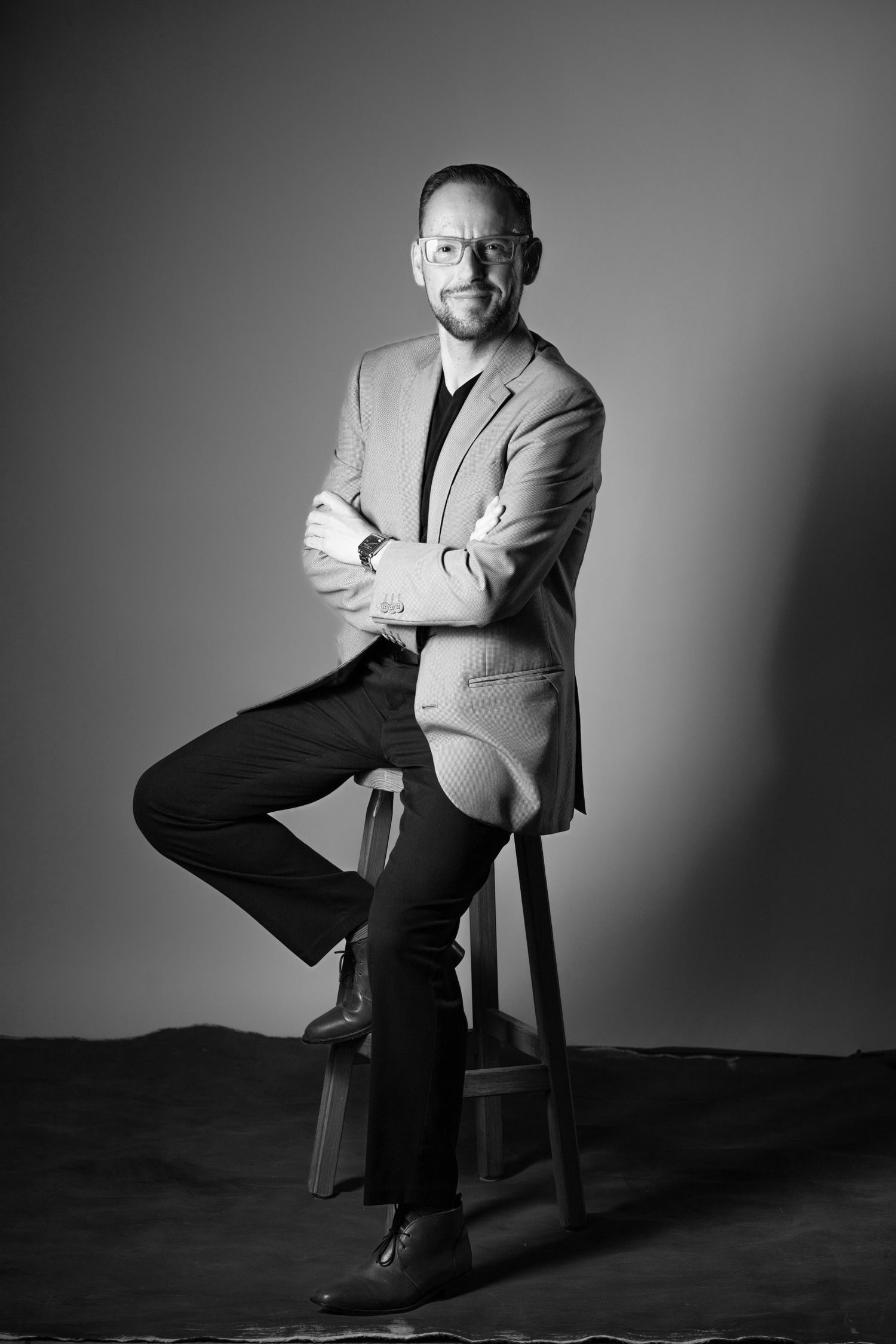 Black and white studio portrait of person in blazer and glasses seated on stool with crossed arms against gray backdrop.