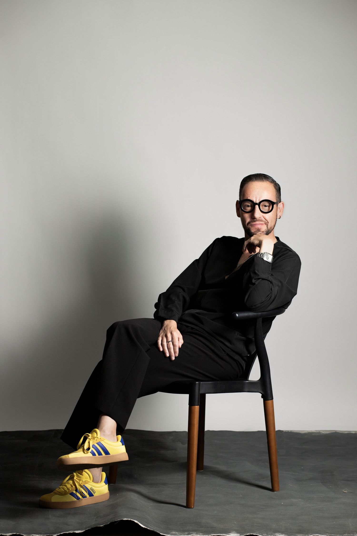 Person in all black attire sits on wooden chair wearing bright yellow sneakers against grey backdrop in artistic poses.