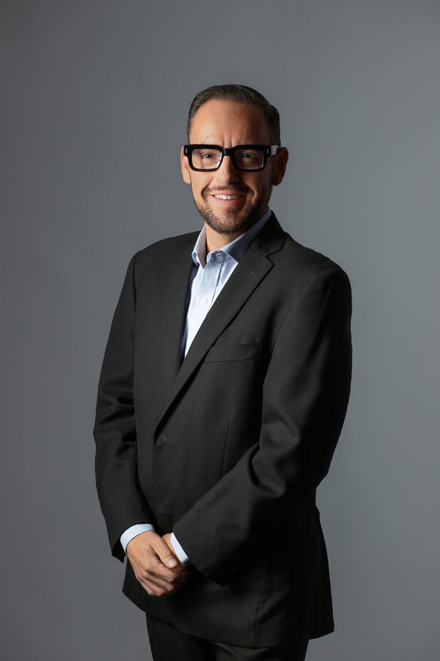 Professional headshot series showing different poses of someone wearing a dark suit and glasses against a gray background.